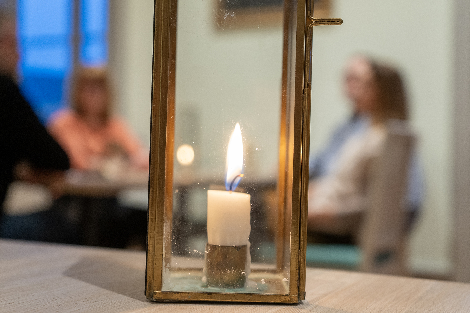 Fyra personer som samtalar vid ett bord syns i bakgrunden, men i fokus närmast betraktaren brnner ett stearinljus i en lite medfaren ljuslykta.