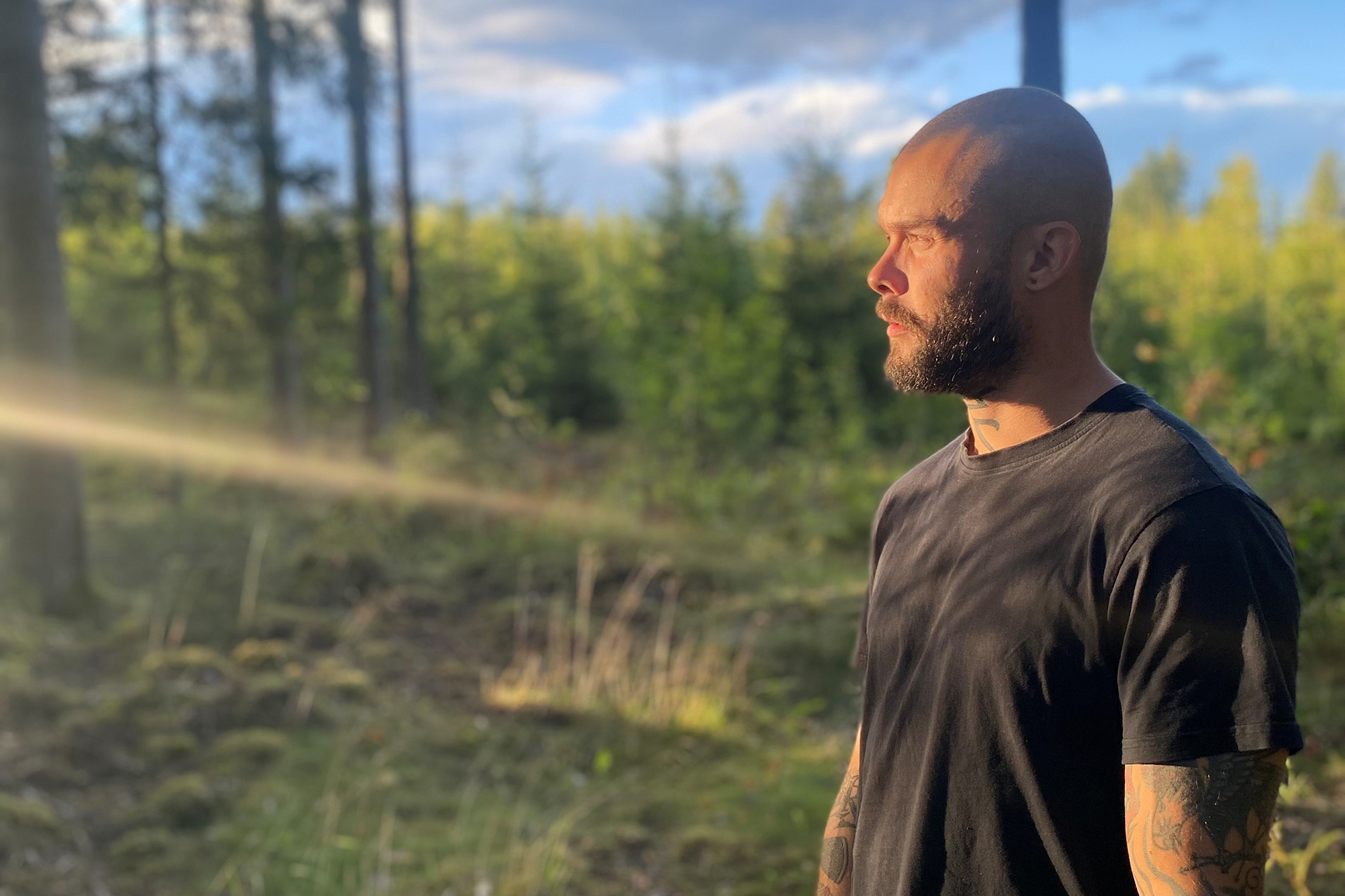 Sångaren Wille Matki, med mörkt skägg, t-shirt och tatuerade armar, står i en ljust upplyst skogsdunge, sedd i profil med solen i ansiktet och blicken fäst på något i fjärran.