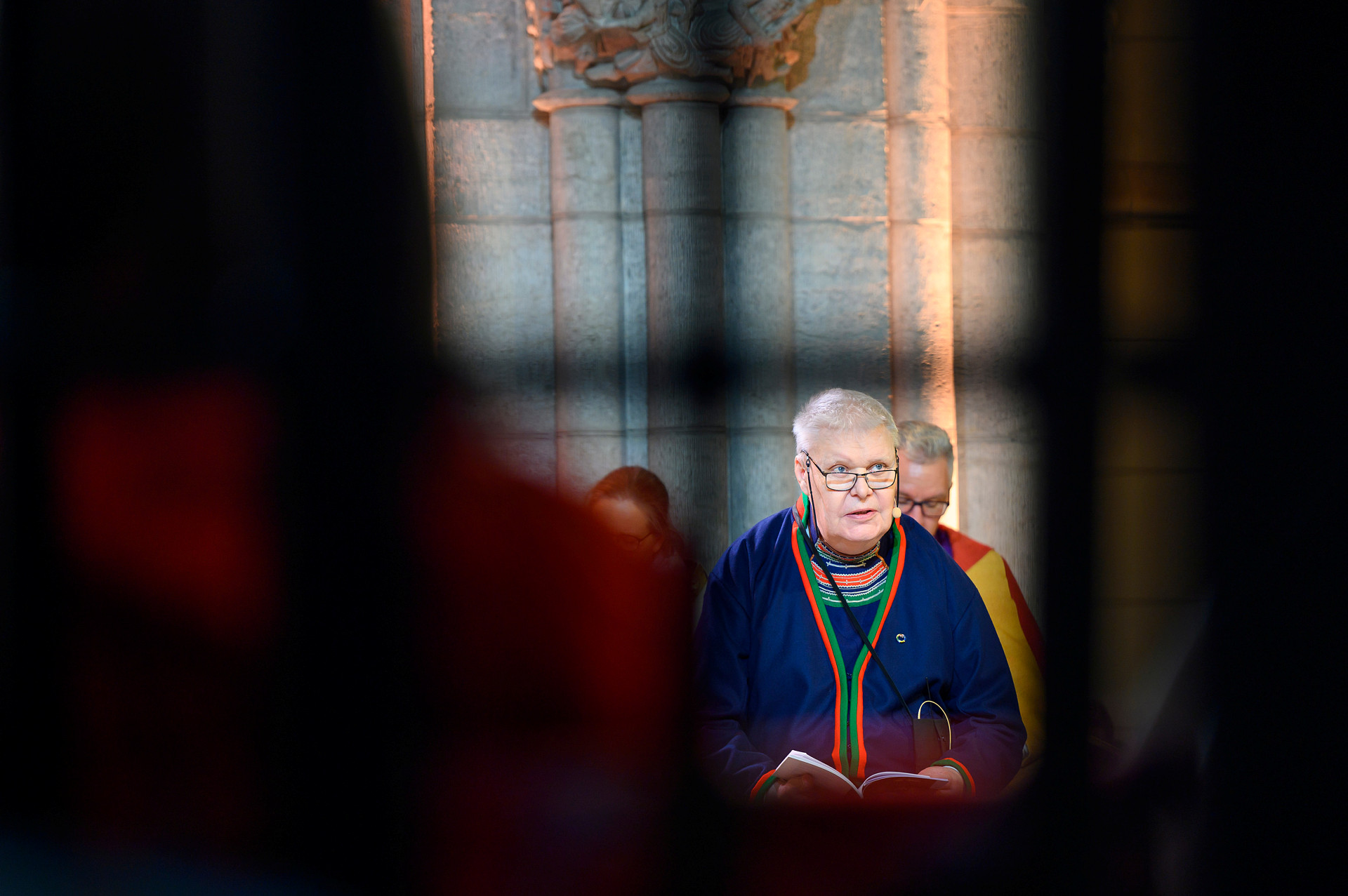 Man, klädd i samisk klädsel, framträder mellan skuggor av andra personer i en kyrka. 
