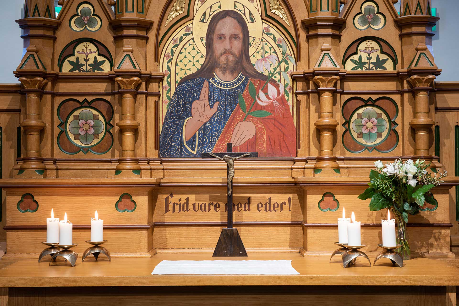 Altare med tända ljus. Altartavla bakom föreställande Jesus med en rosa lilja och handen höjd i välsignelse.