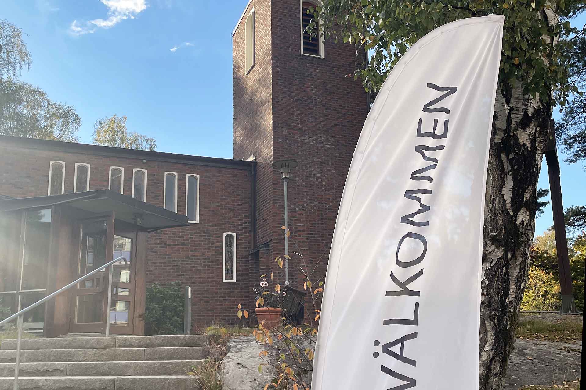 En flagga med texten "Välkommen" framför en kyrka i tegel en sommardag.