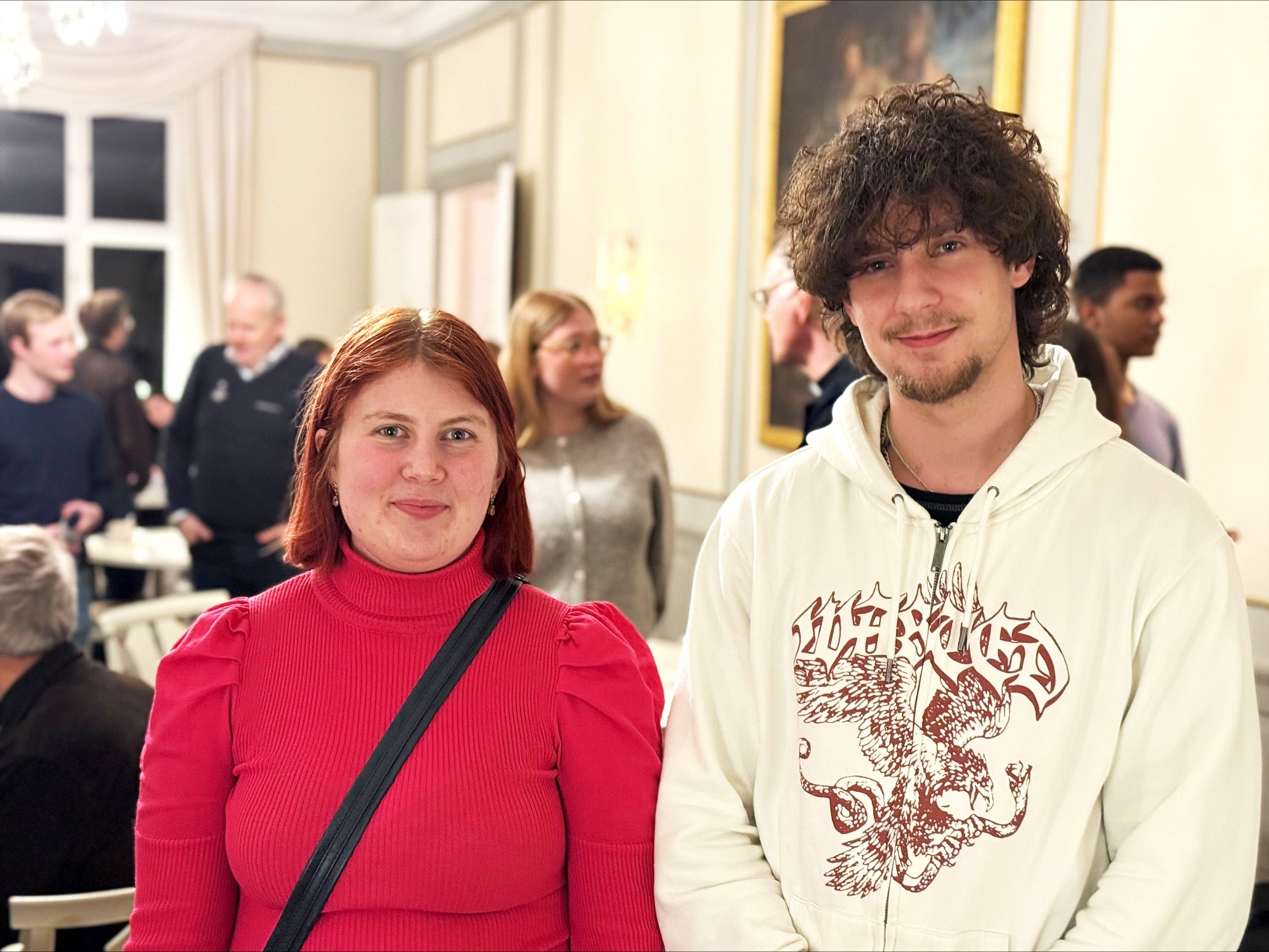 Bild i halvfigur, till vänster står Majken i röd tröja, till höger står Edisson i vit hoodie. I bakgrunden står andra deltagare under Nyfiken på-kvällen och samtalar med varandra.