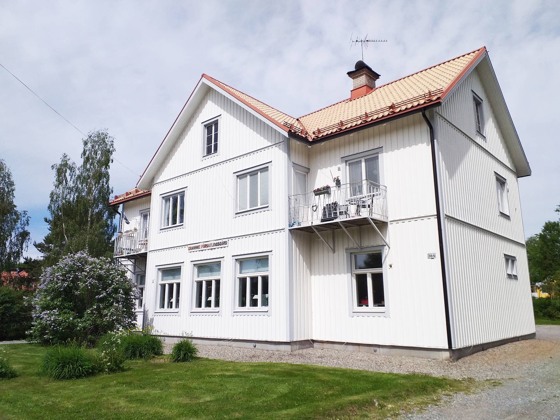 Graninge församlingsgård. En vit träbyggnad. På gräsmattan utanför blommar en syren.