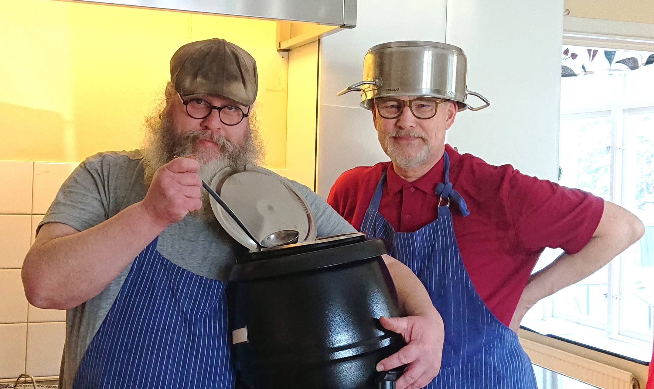 Två män i randiga köksförkläden. Den ene håller i en soppkittel och soppslev. Den andre har en stor kastrull på huvudet.