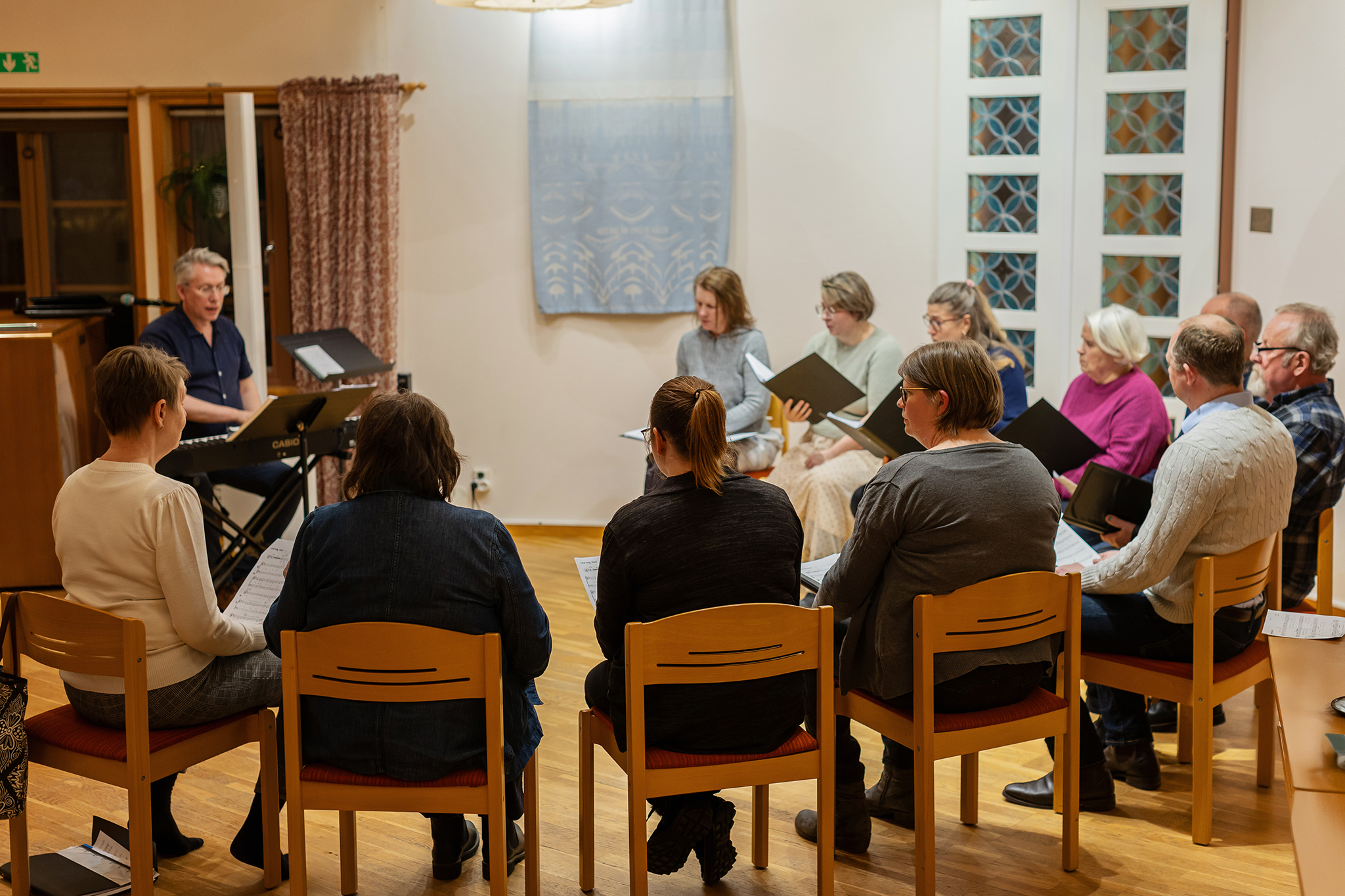 En grupp körsångare som sitter i en halvcirkel på stolar med ryggen mot kameran. 