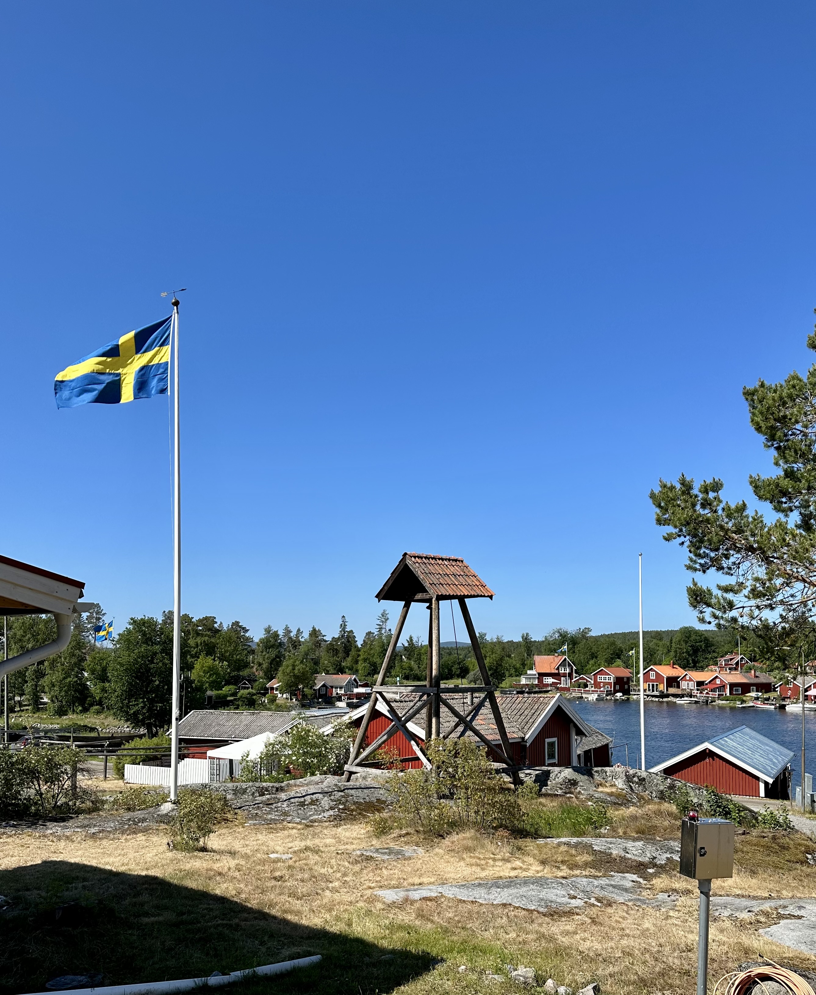 Sommar i en skärgårdsidyll i strålande sol. Röda fiskebodar, en svenska flagga blåser i vinden och ett klocktorn i trä är i blickfånget.