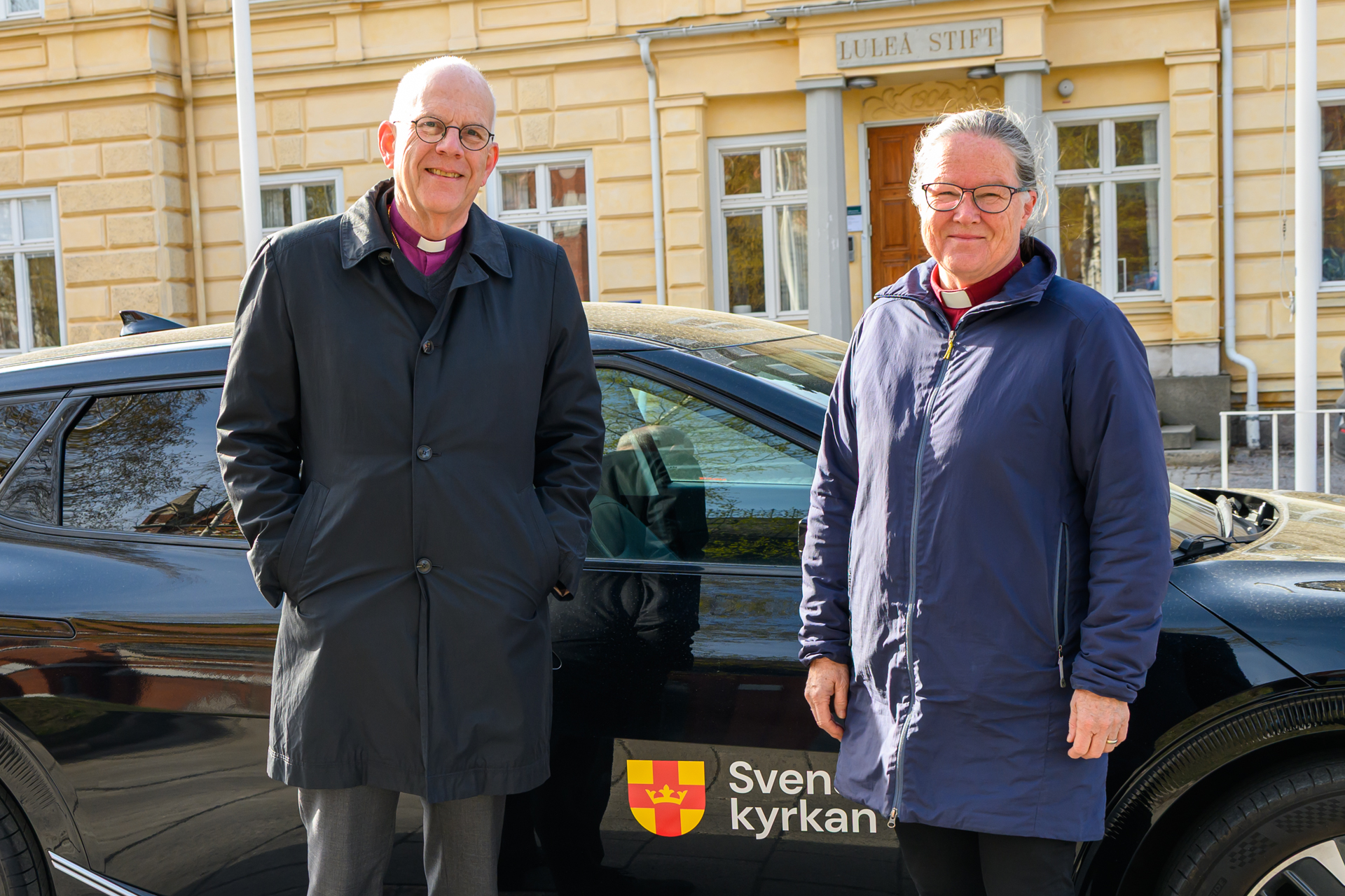 Två personer står framför en bildörr med Svenska kyrkans logotyp på.