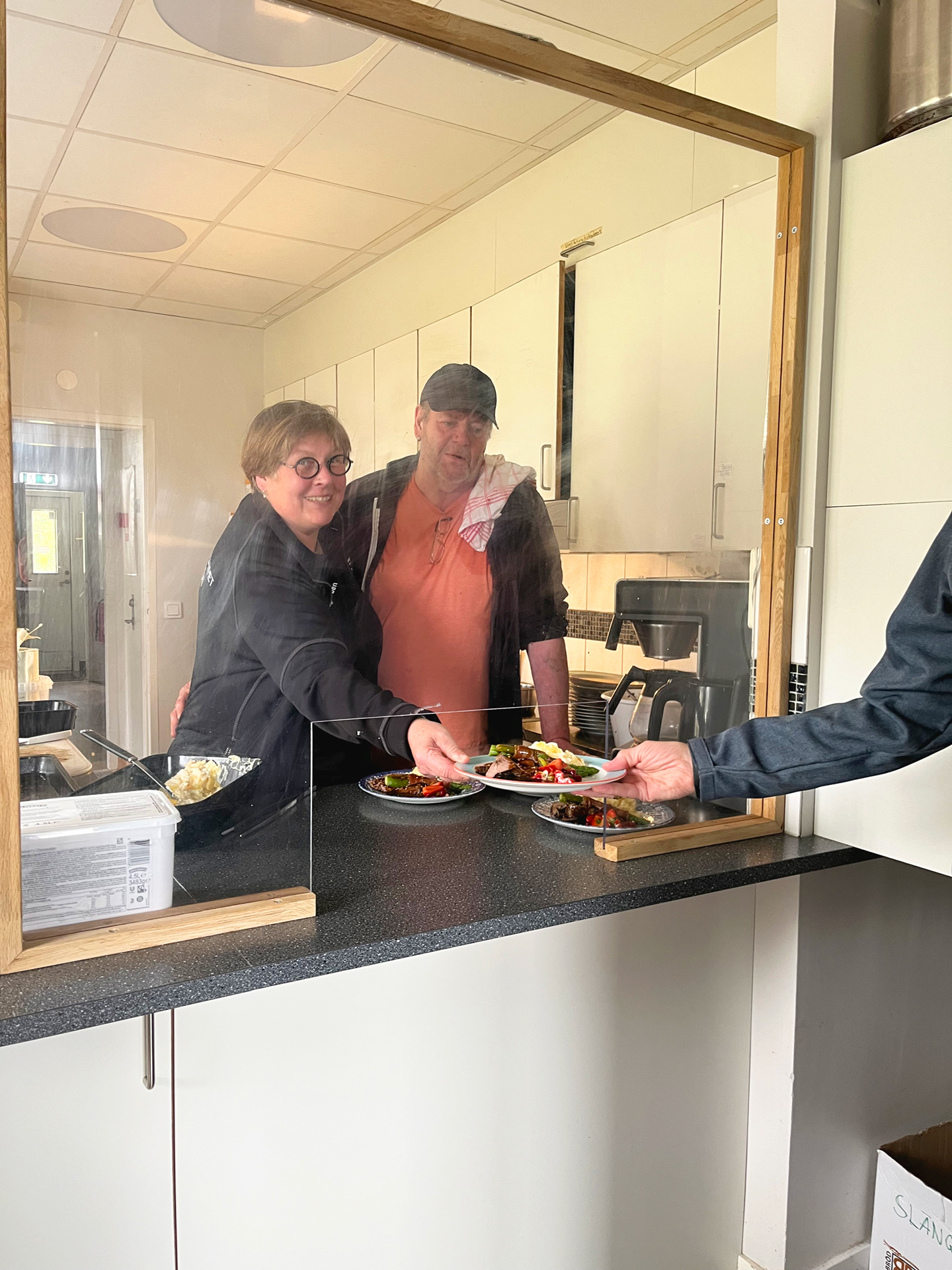 Två personer serverar mat ur en serveringslucka.