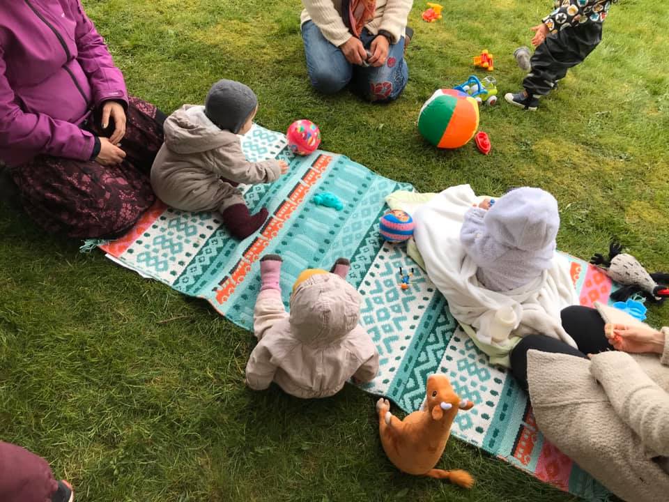 Barn och föräldrar sitter på en färgglad matta utomhus och leker. 