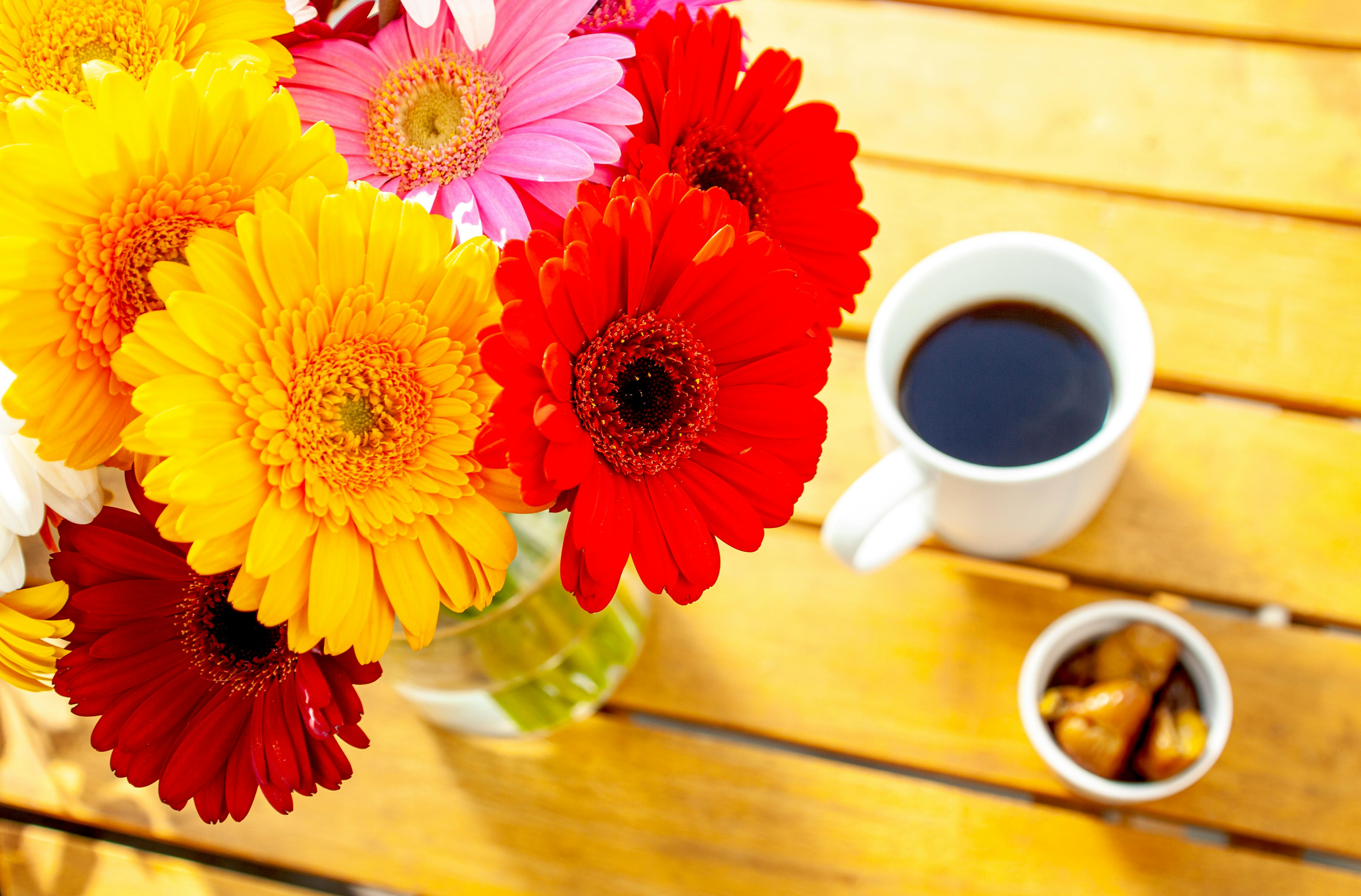Färgglada blommor i en vas tillsammans med en kaffekopp på ett gult bord.
