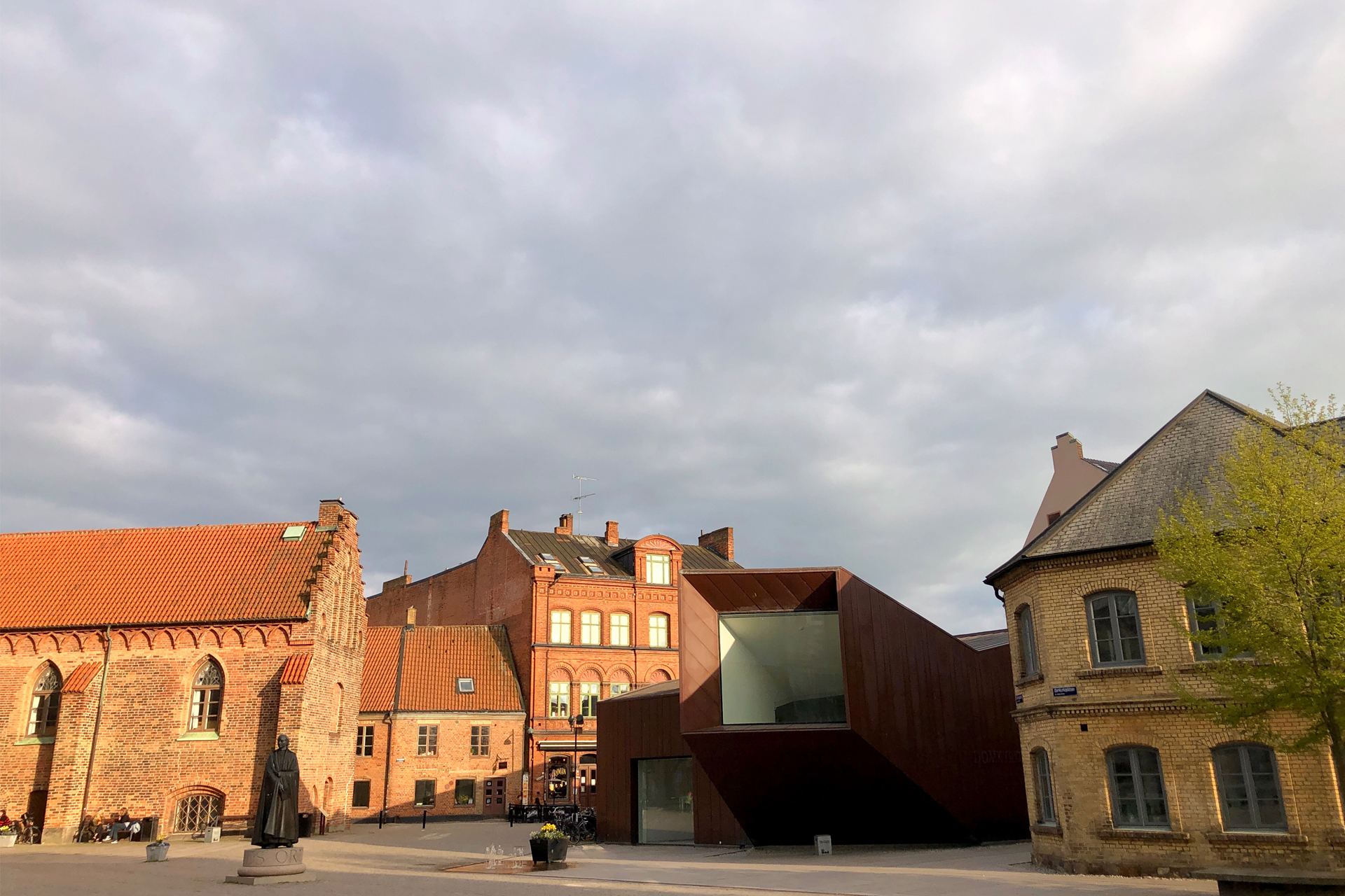 Ett torg med två röda tegelbyggnader, en modern byggnad i corten och en gul tegelbyggnad. På torget står en staty och det finns en fontän.