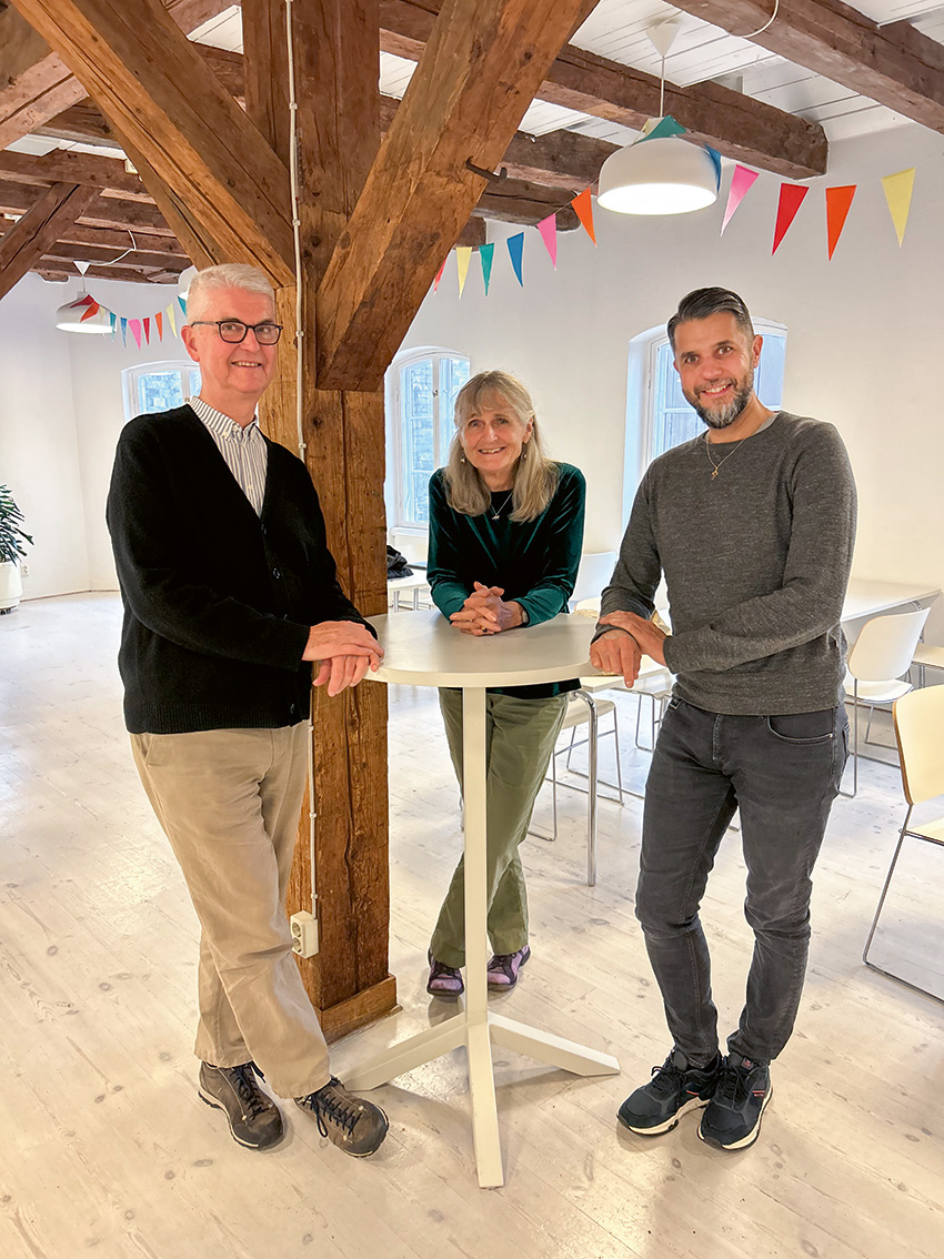 Tre personer som står runt ett litet bord mot en fond av gamla bjälkar.