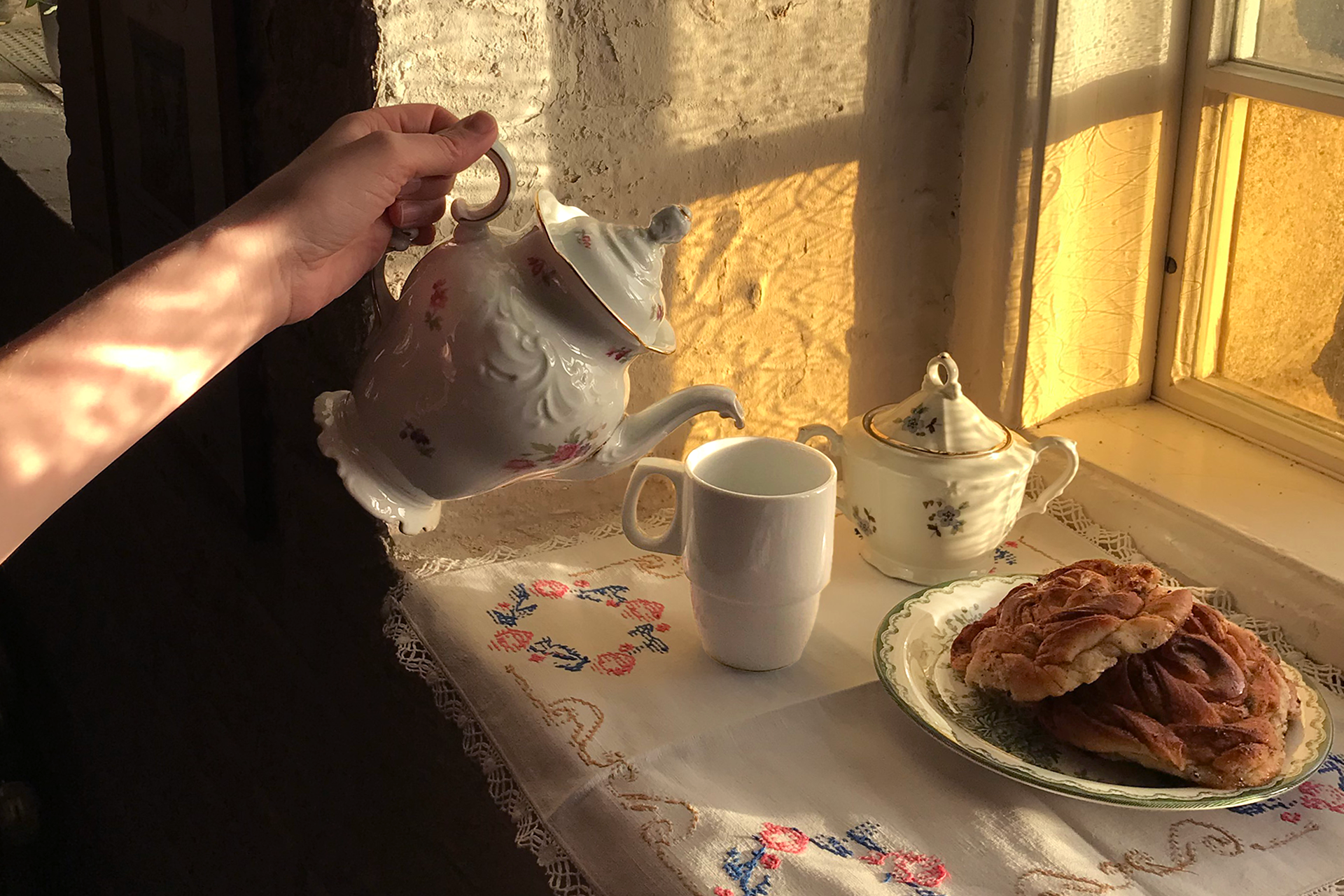 Någon häller upp te från en gammal engelsk tekanna i en tekopp. Bredvid står ett fat med kanelbullar.