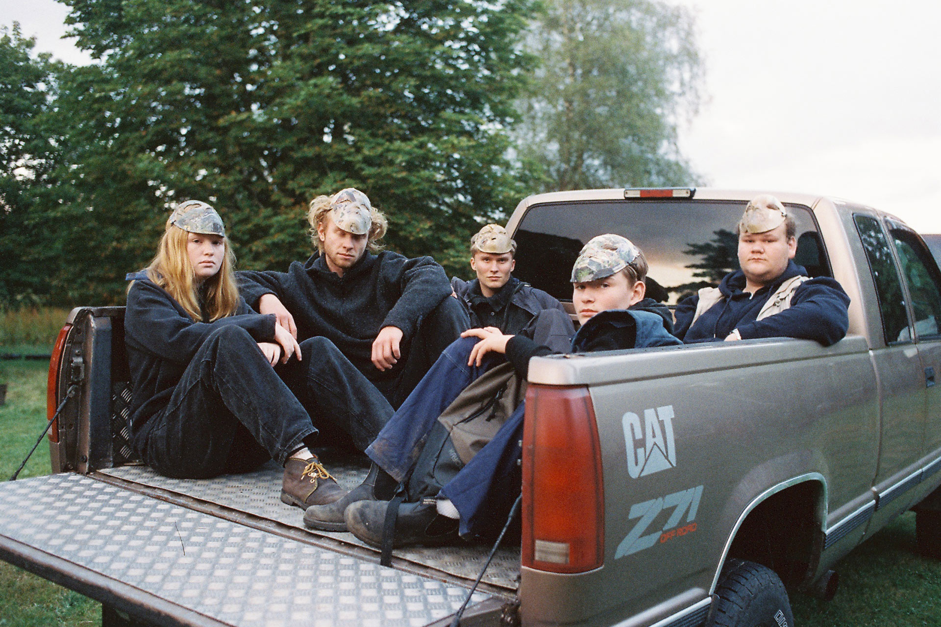 Fem ungdomar sitter på flaket på en bil. De är klädda i blå overaller och har masker på huvudet.