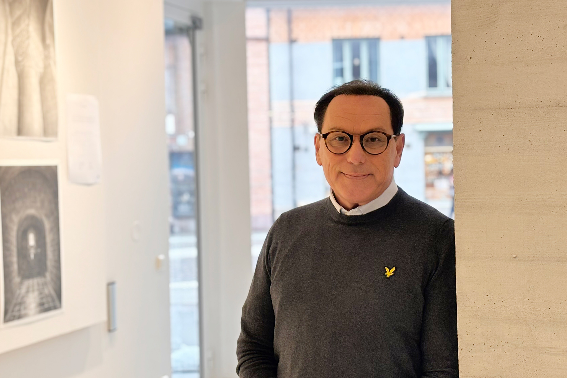 En man med mörkt hår och glasögon står lutad mot en vägg. 