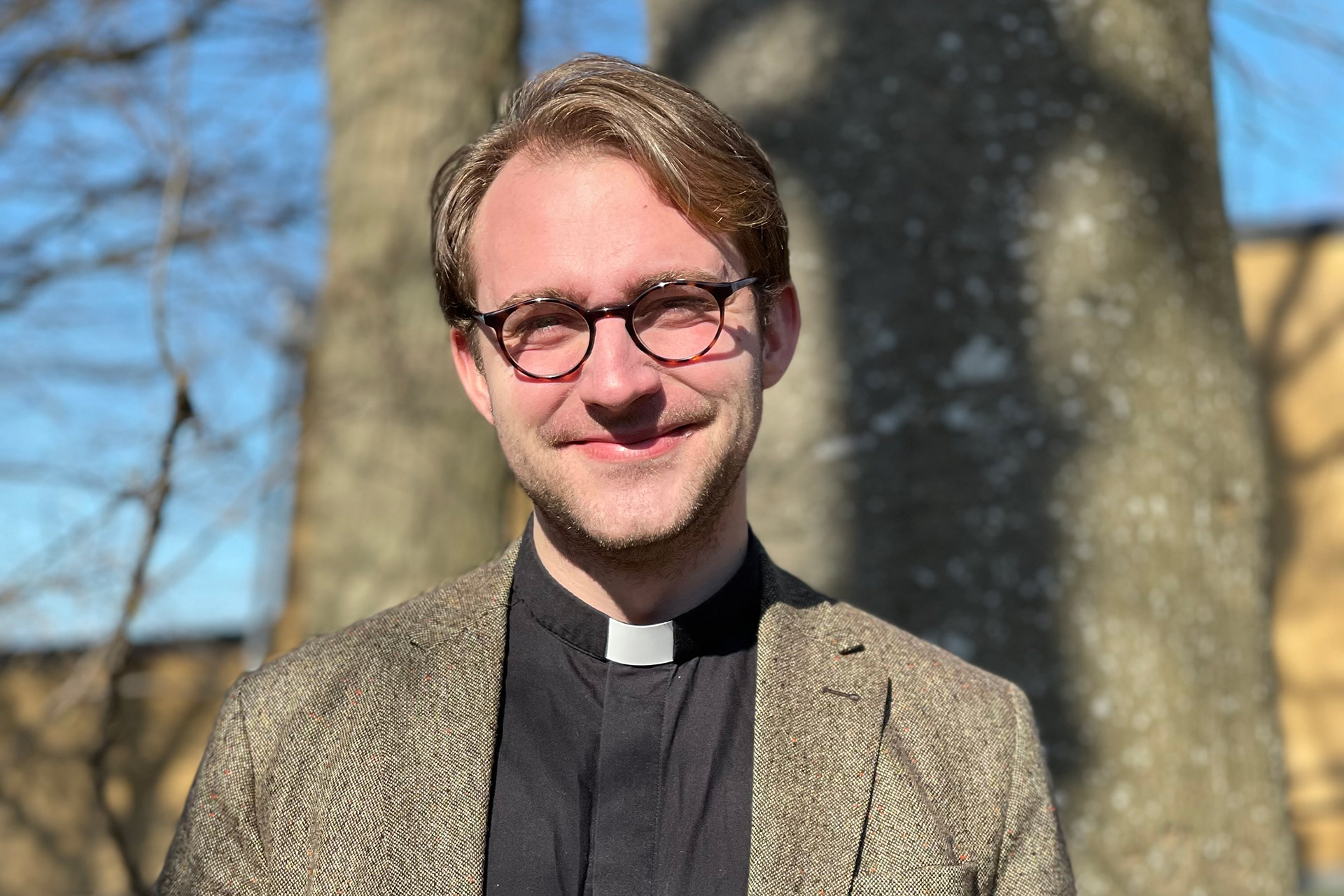 En ung smal man klädd i svart prästskjorta och brunmelerad kavaj. Han har runda glasögon och lätt skäggstubb. I bakgrunden syns tjocka trädstammar och kala grenar mot en blå himmel och en gul fyrkantig tegelbyggnad.