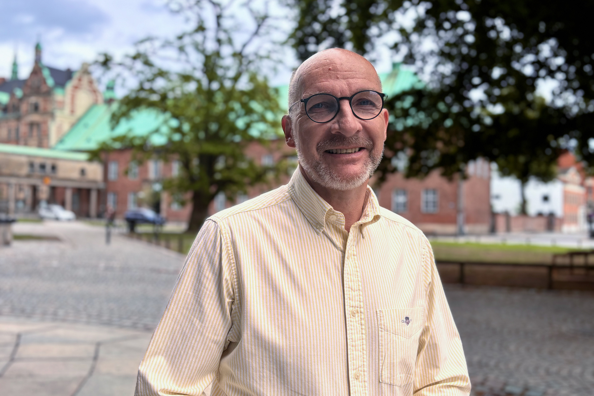 Man med skalligt huvud och glasögon, klädd i randig vit skjorta. I bakgrunden grönskande träd och stadsmiljö.