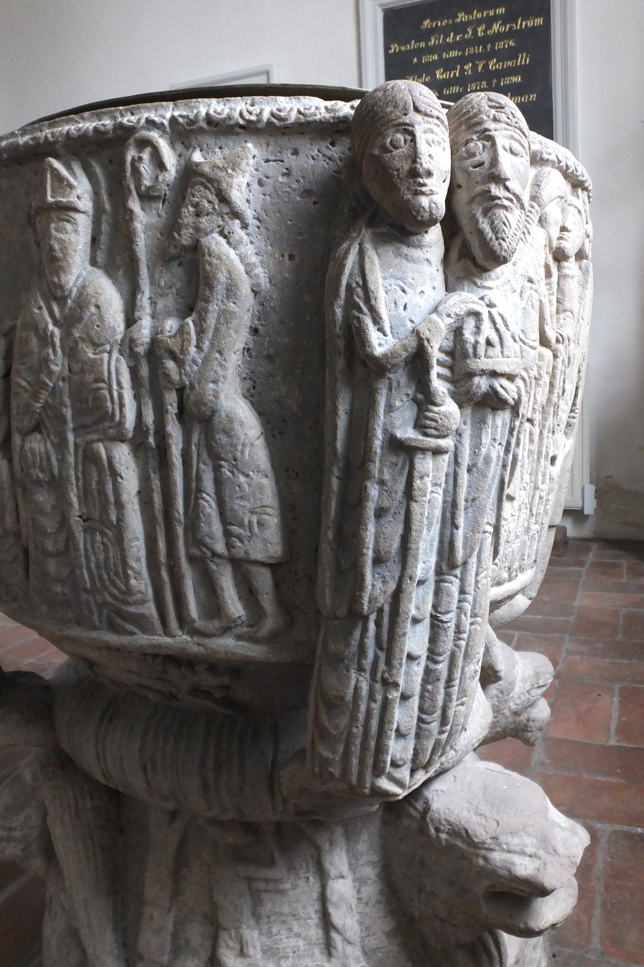 Dopfunten i sten i Tryde kyrka har uthuggna bilder. Bilderna föreställer grupper av människor.
