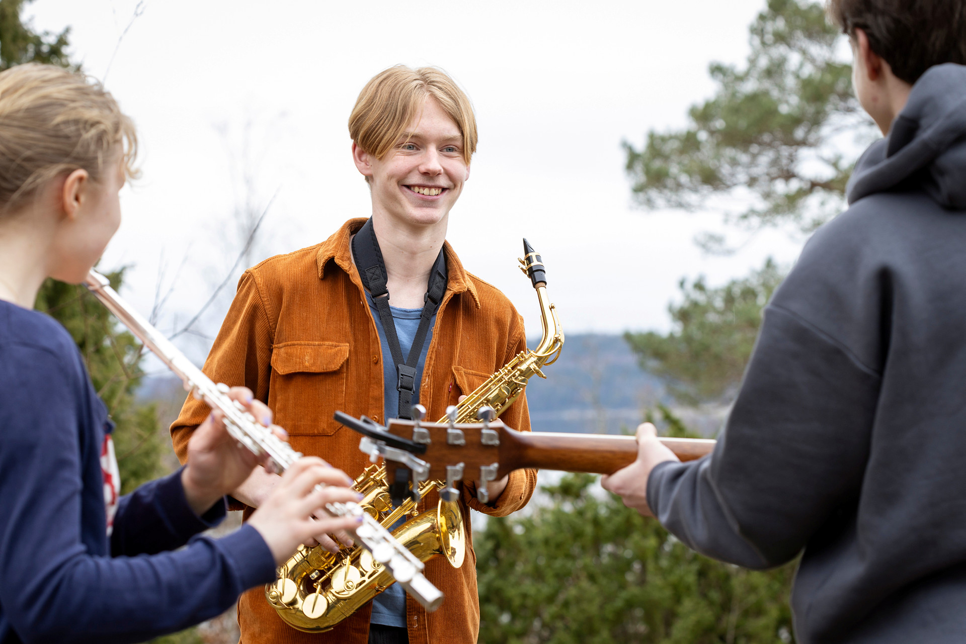En ung kille håller i en saxofon och ler mot vänner, som spelar på en gitarr och en flöjt.