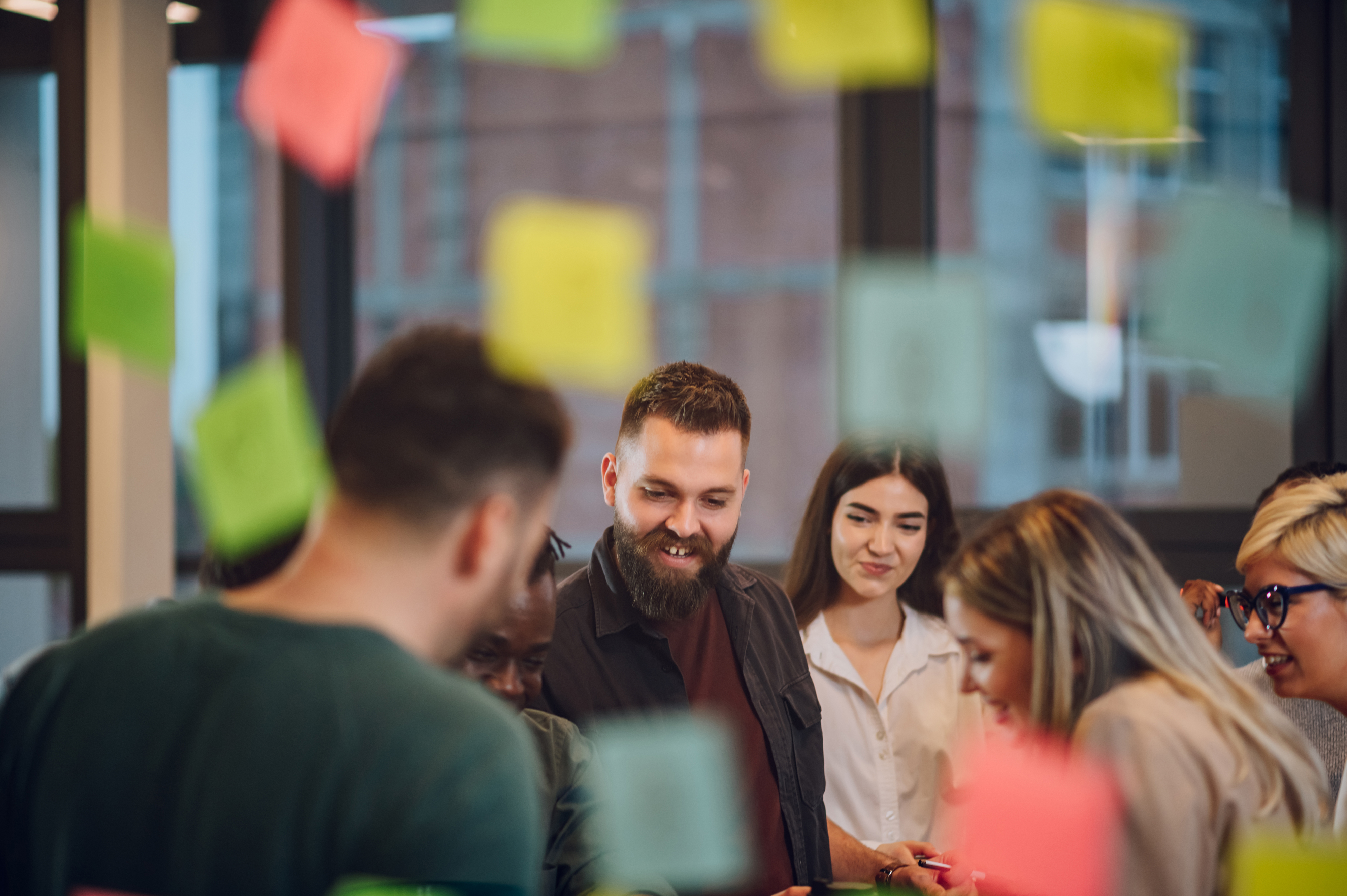 I förgrunden hänger post-it-lappar och bakom syns en grupp människor som samarbetar och ler.