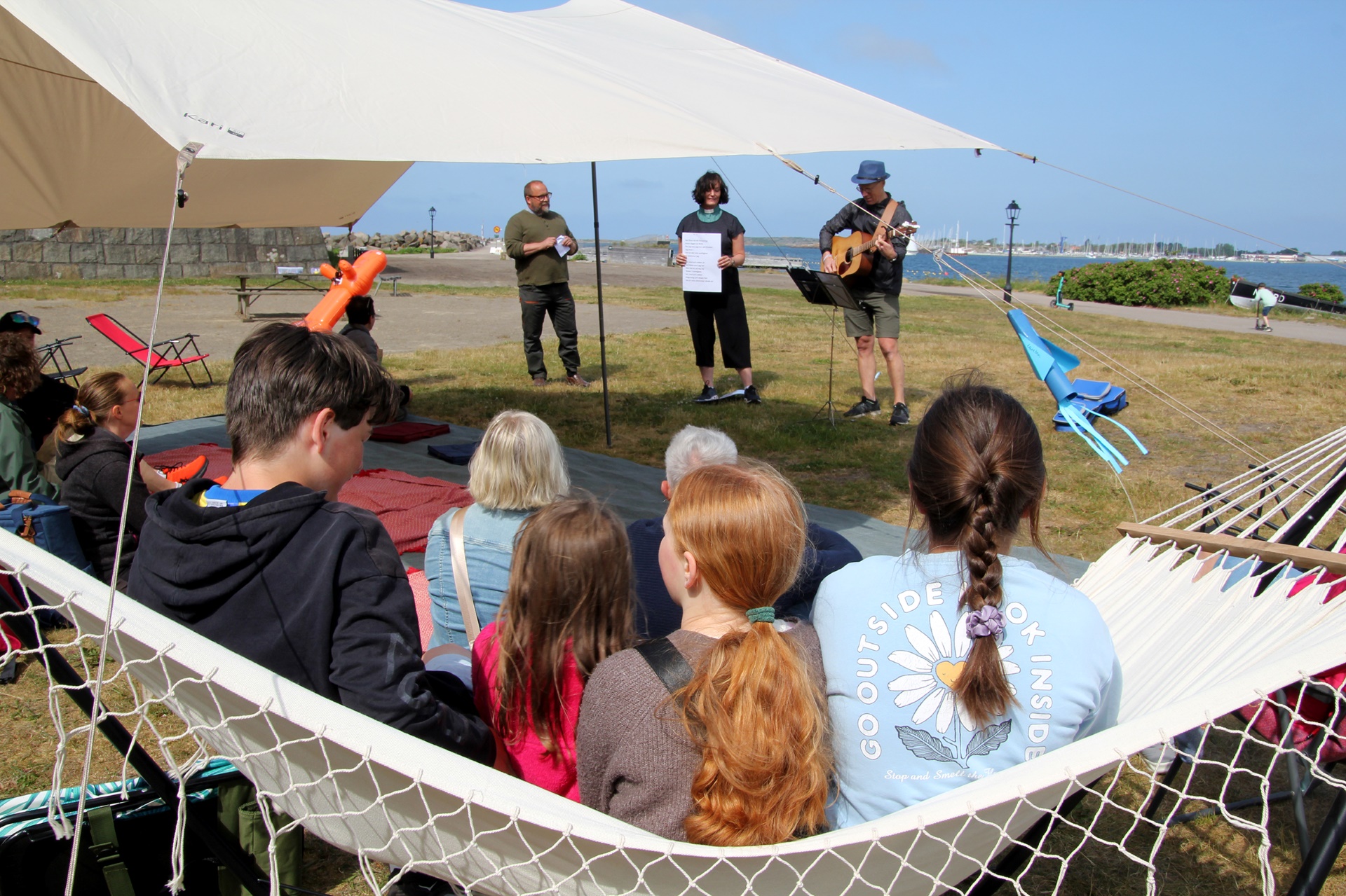 Fyra personer sitter i en hängmatta. Framför dem sitter fler personer och längst fram står tre personer som leder gudstjänsten.