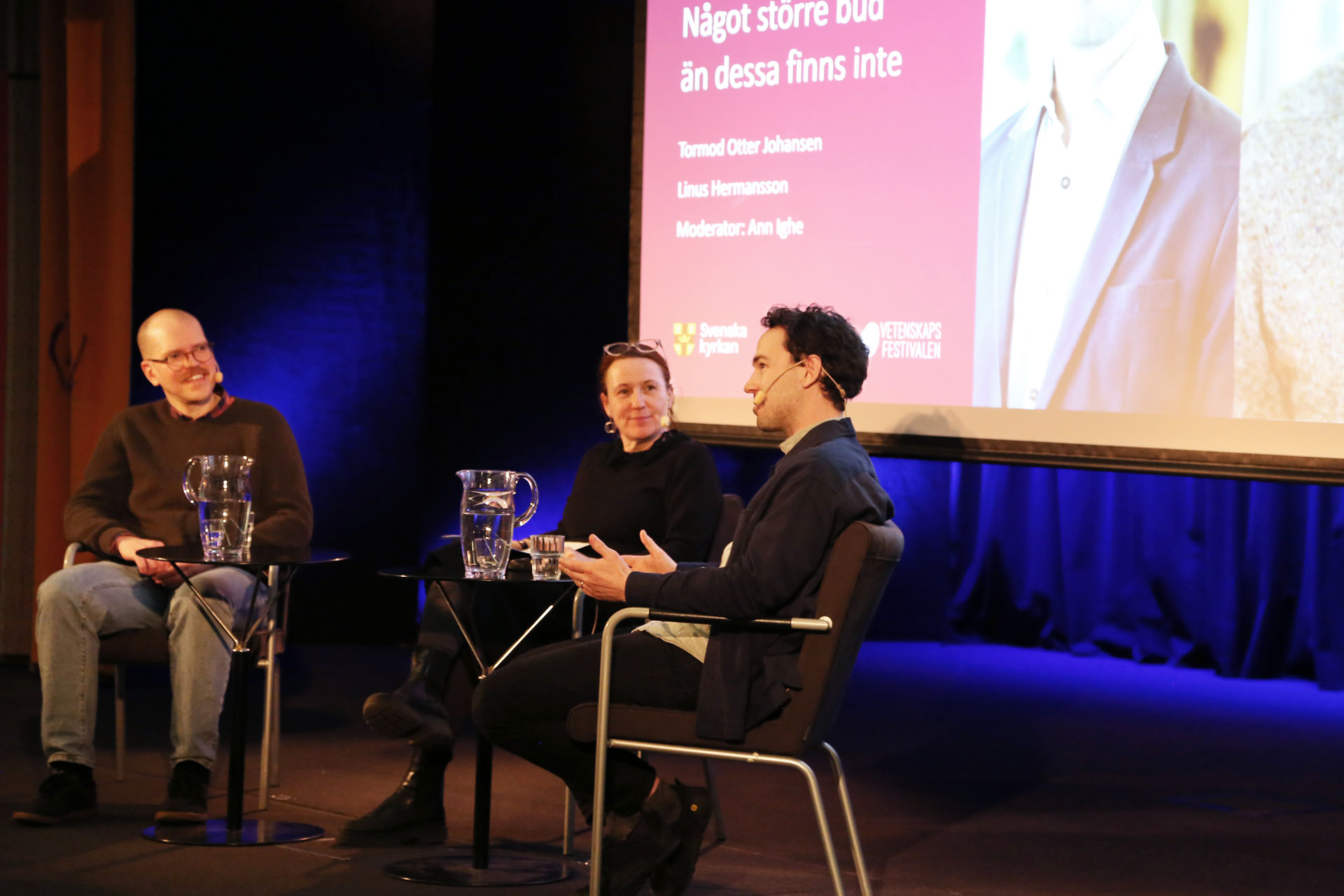 Tre personer sitter på en scen. En talar och gestikulerar och de andra lyssnar med glada miner. 