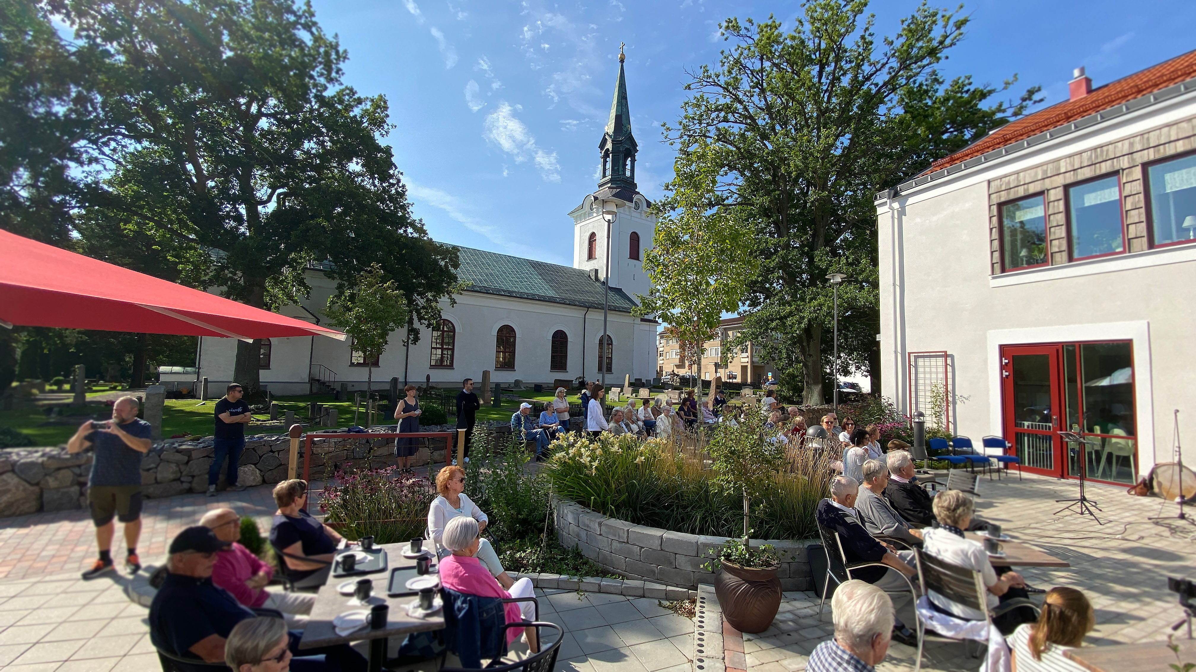 Människor sitter i solen och lyssnar på livemusik, i bakgrunden syns tornet på en kyrka.