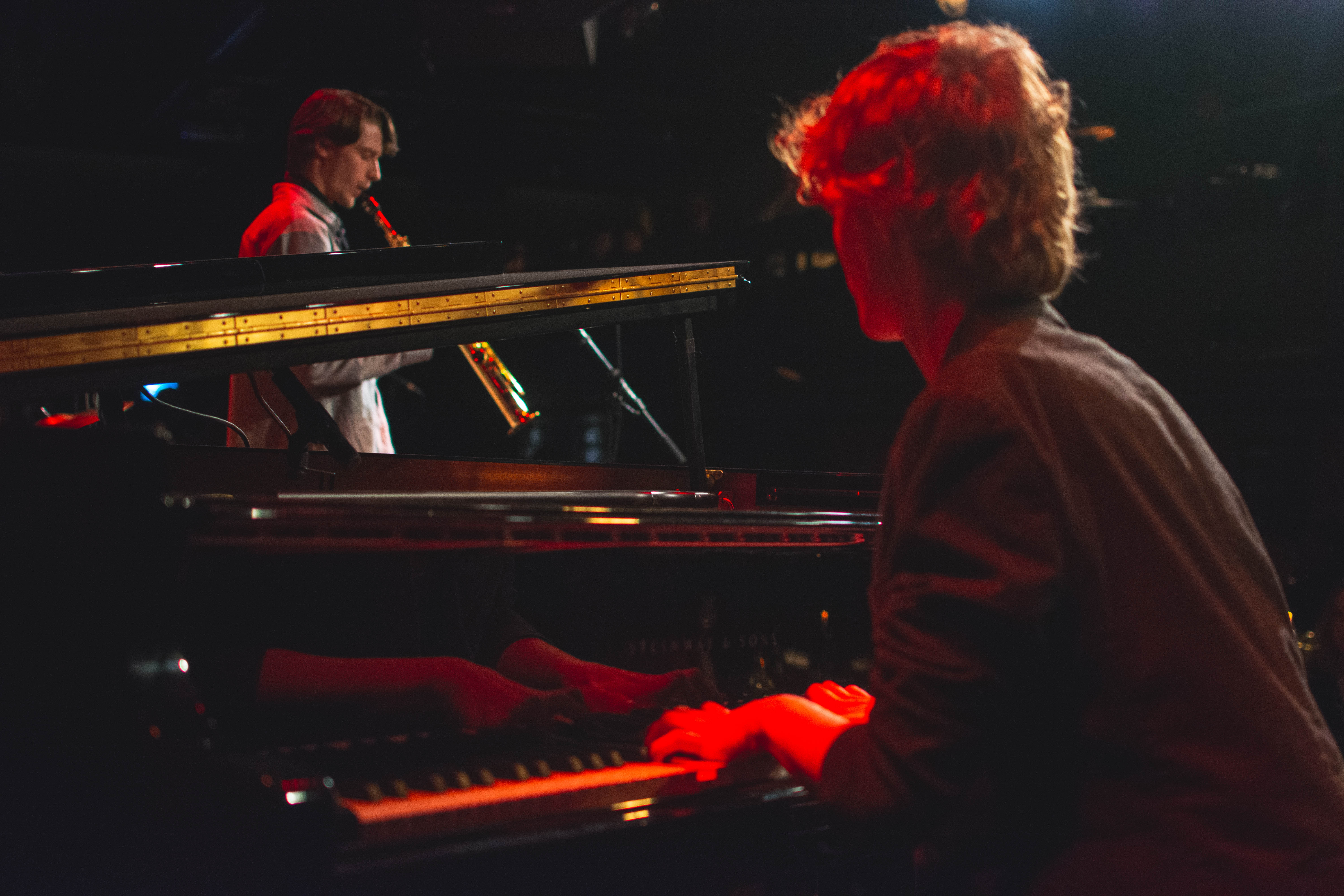 Jazzduon Melker Falk och Joel Wetsreling syns musicera på piano och saxofon i ett stämningsfullt foto med dovt röda och svarta toner.