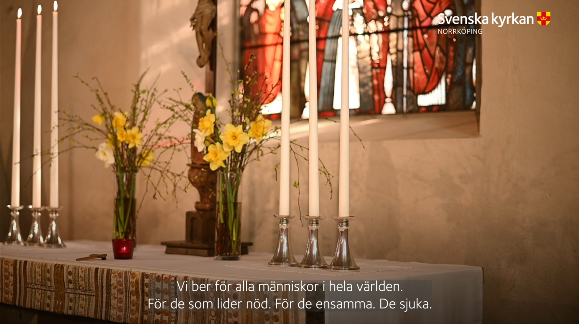 Ett altare med påskliljor, brjörkris, krucifix och sex stycken höga altarljus. I bakgrunden anas det färgglada fönstret i koret.