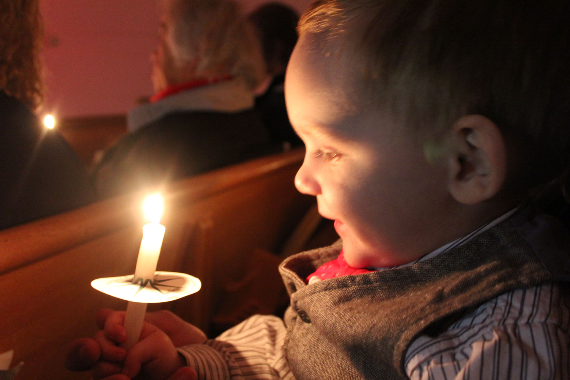 Ett leende litet barn tittar in i ett ljus sittandes i en kyrkbänk. I bakgrunden syns ryggar på andra personer. 