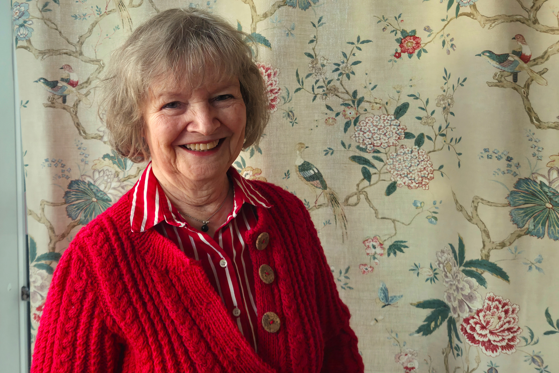 En glad kvinna står i rödvit blus och röd kofta framför en blommig gardin.