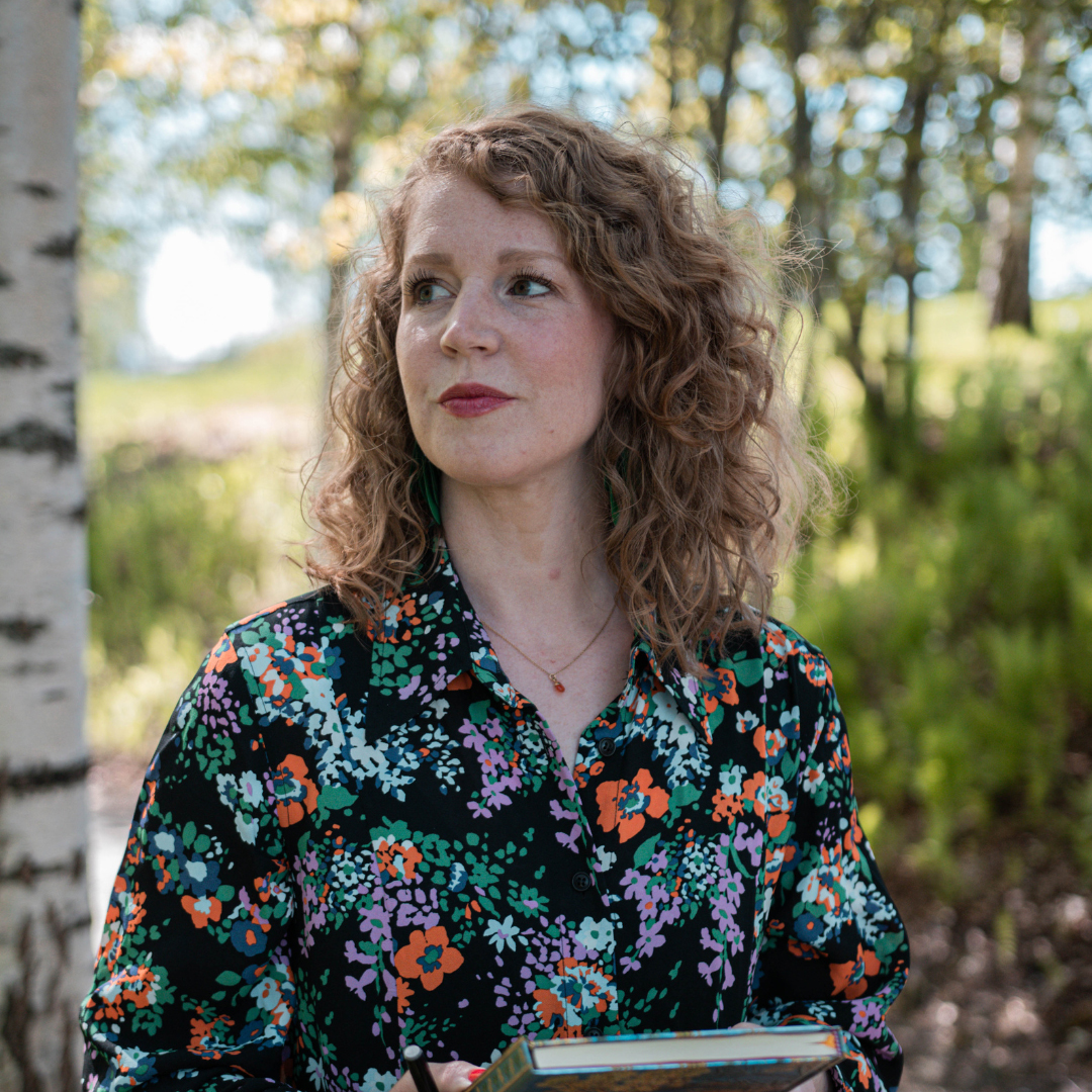 Porträttbild på Elin Malmberg i somrig lövskog. Elin tittar åt sidan och håller en bok.