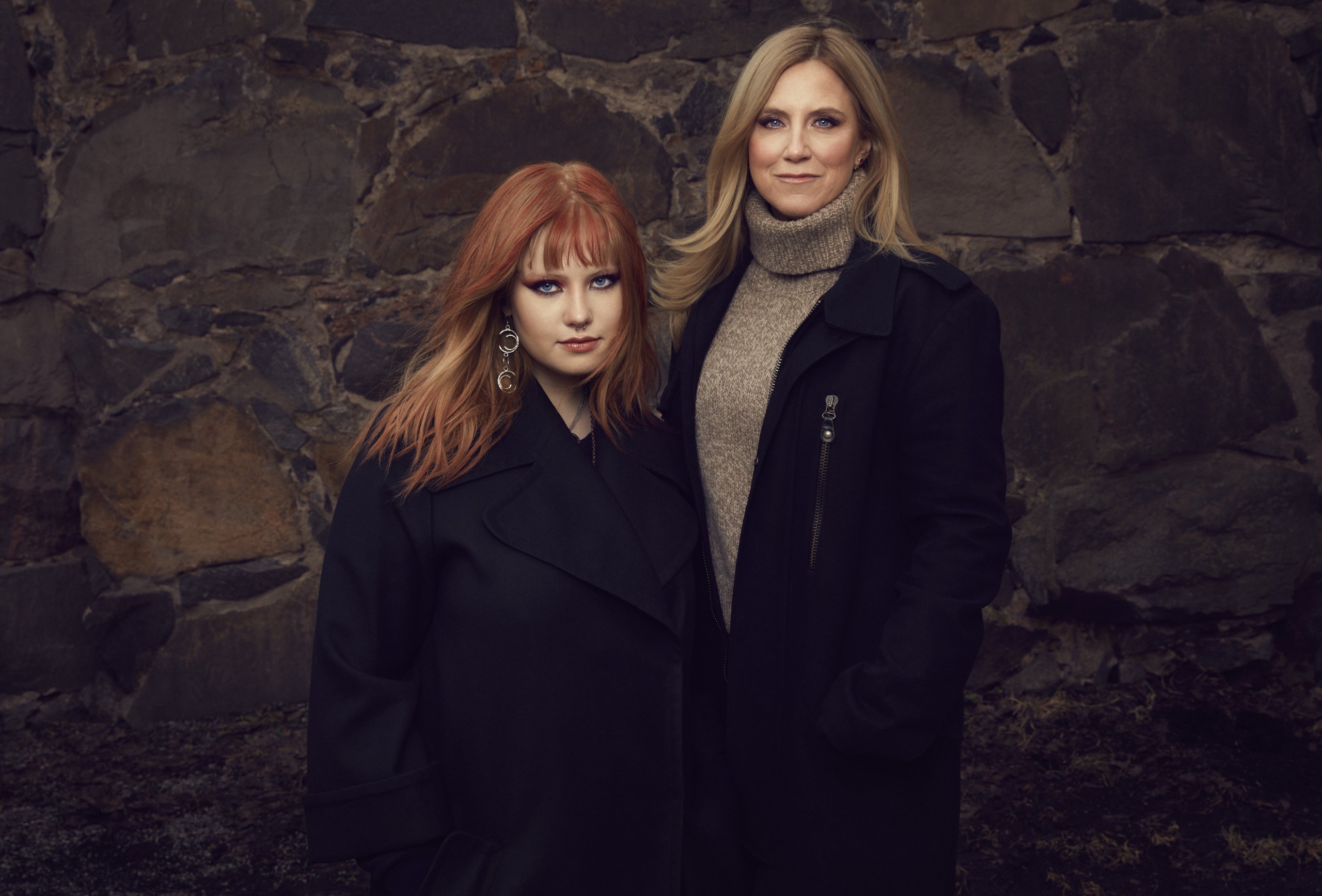 Frida och Tilda Boisen står bredvid varandra framför en grov stenmur utomhus. Båda bär mörka ytterplagg; personen till vänster har kopparrött hår med lugg och stora hängande örhängen, medan personen till höger har ljusare, längre hår och en stickad polotröja under en mörk kappa. Bilden är ett uppställt porträtt med mjukt ljus och dämpade färger.