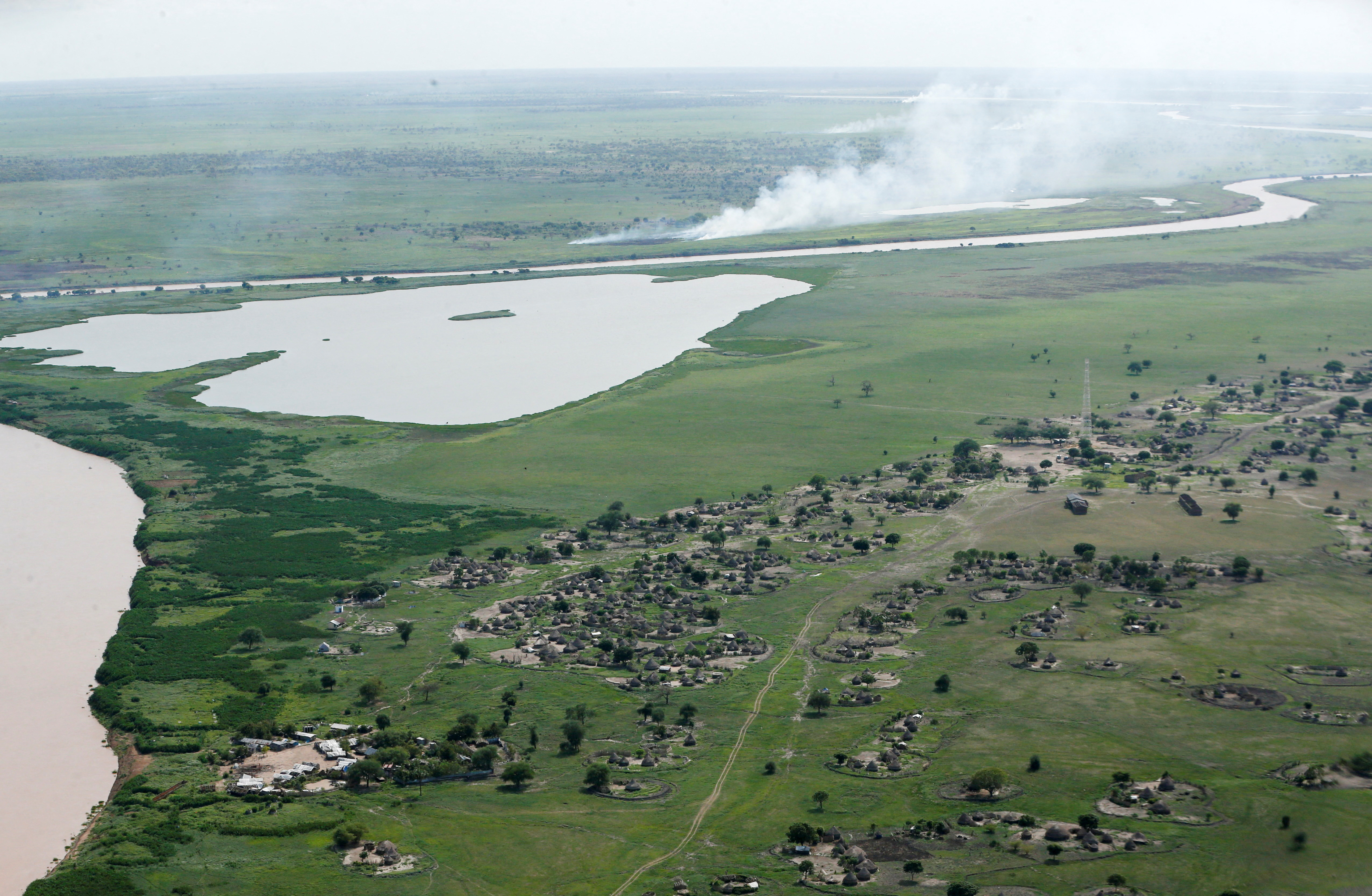 Platt grönt landskap med by och en flod och rök som stiger på avstånd.