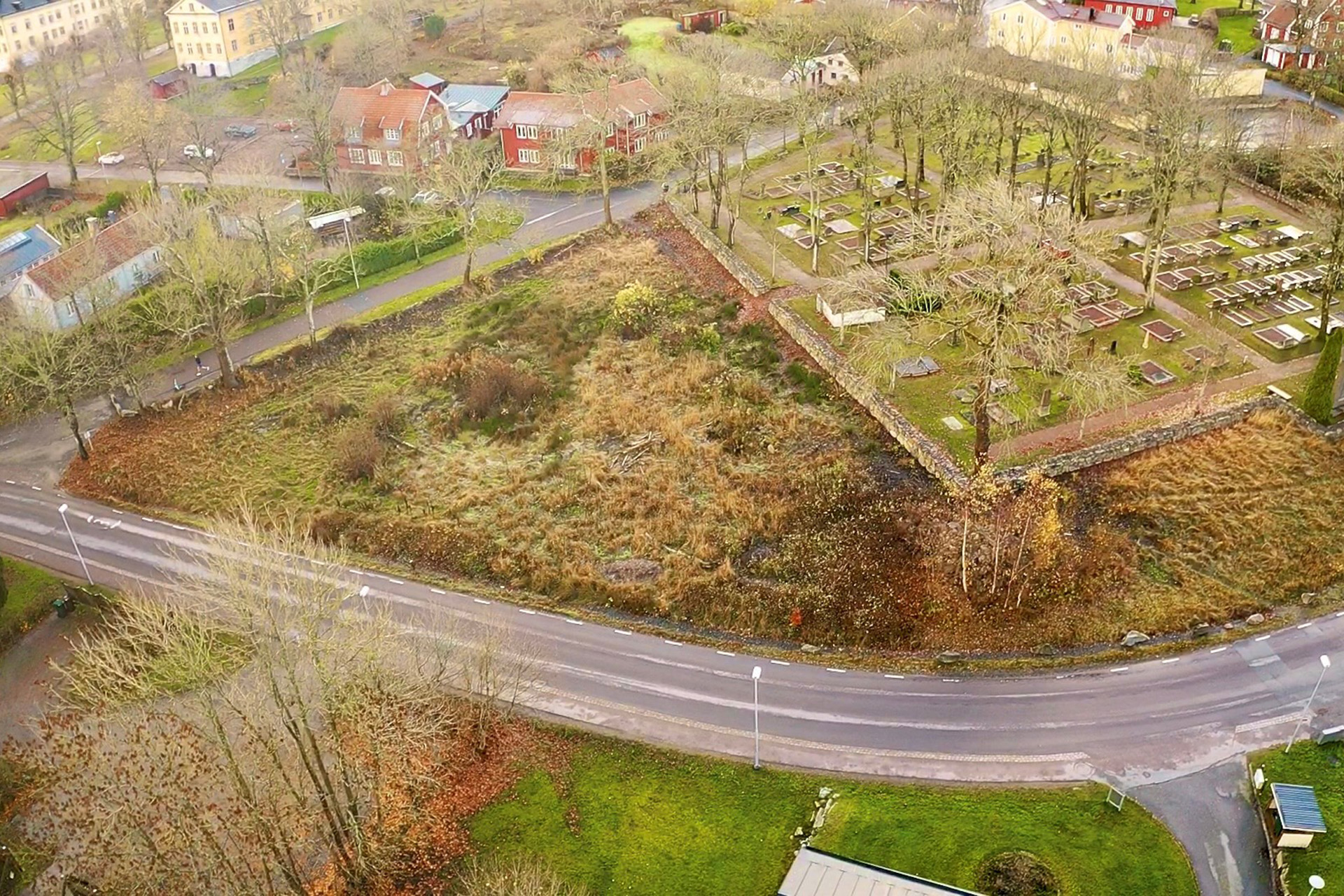 Drönarbild visar en kyrkogård med en markyta intill, träd och hus i bakgrunden.