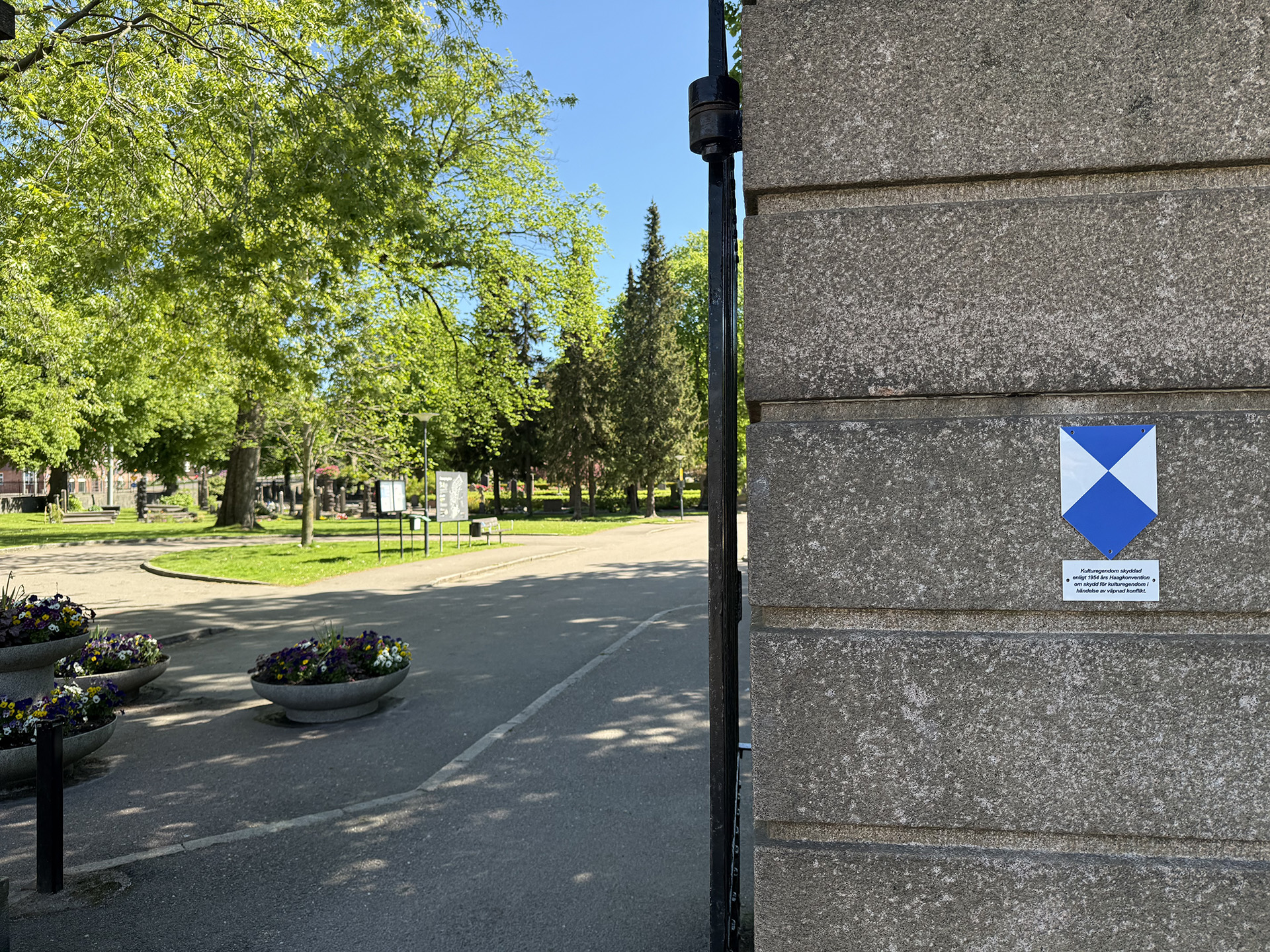 På en stolpe av sten sitter en blåvit sköld, vid sidan av stolpen ser man in på kyrkogården med träd, asfalterad väg och stor kruka.