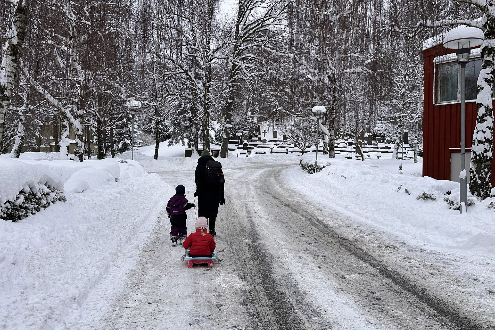 Förälder och barn drar en pulka med ytterligare ett barn i på snötäckt gata i kyrkogårdsmiljö.