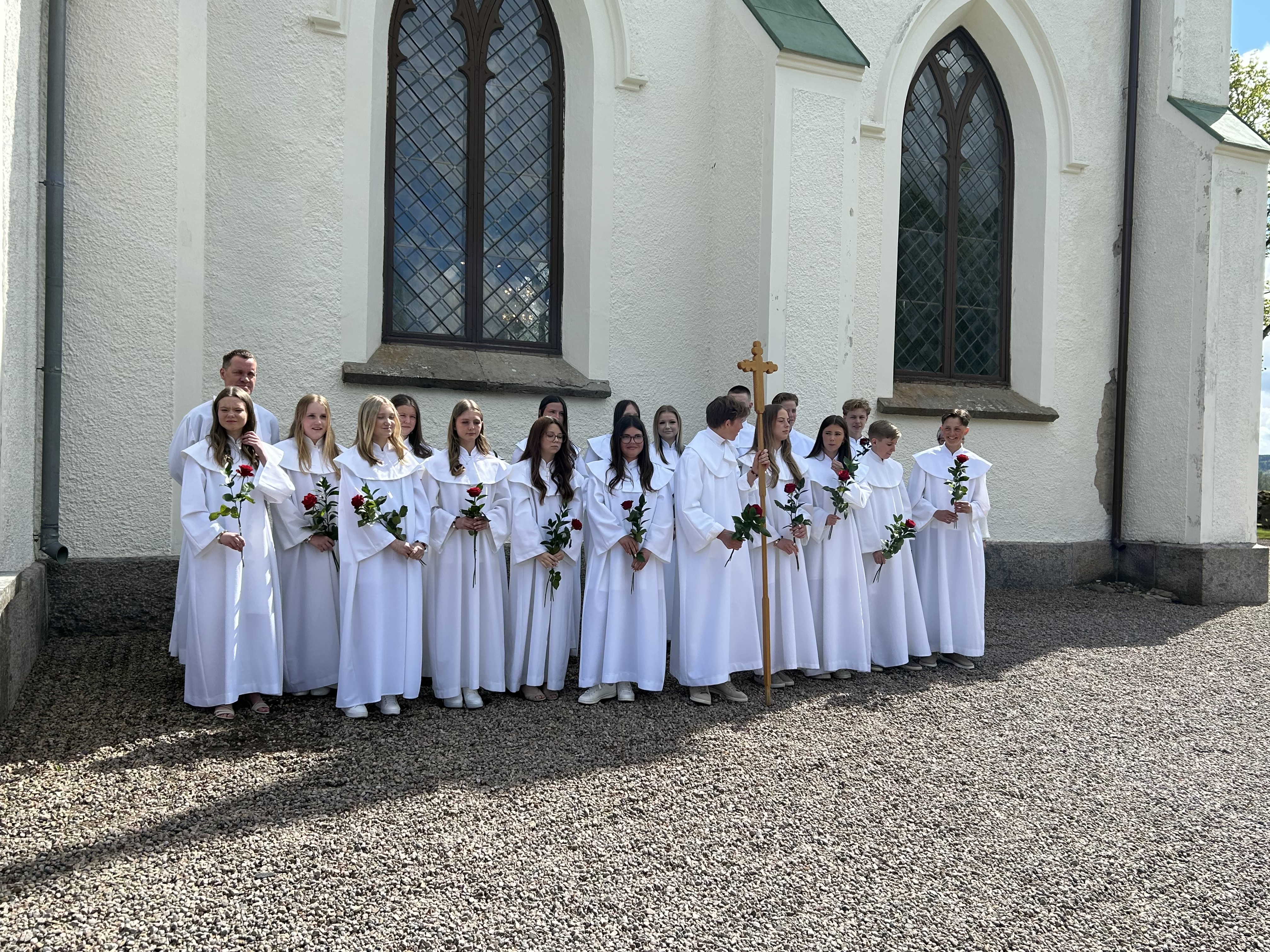 15-20 tjejer och killar i vita kåpor (klänningar) står uppställda på två rader utanför en kyrka.