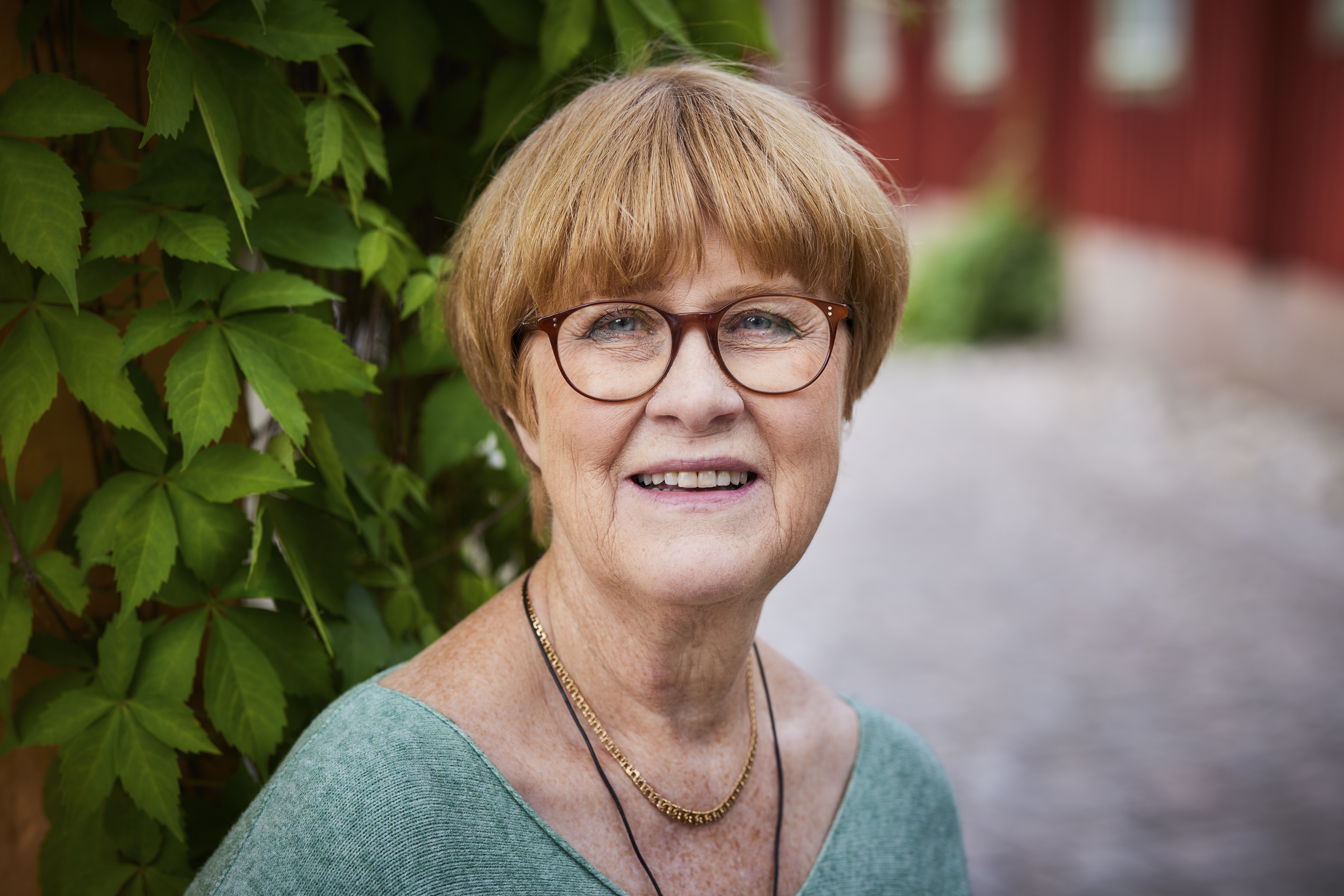 En leende kvinna i 70-årsåldern med kortklippt rödblont hår står framför en buske och en röd vägg.