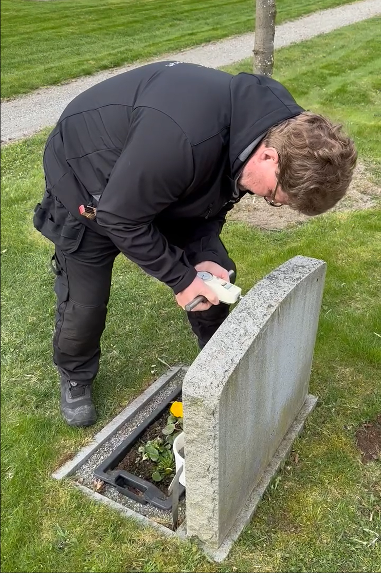 En man i svart jacka utför en gravstenssäkring, genom att trycka en tryckmätare mot en gravsten.