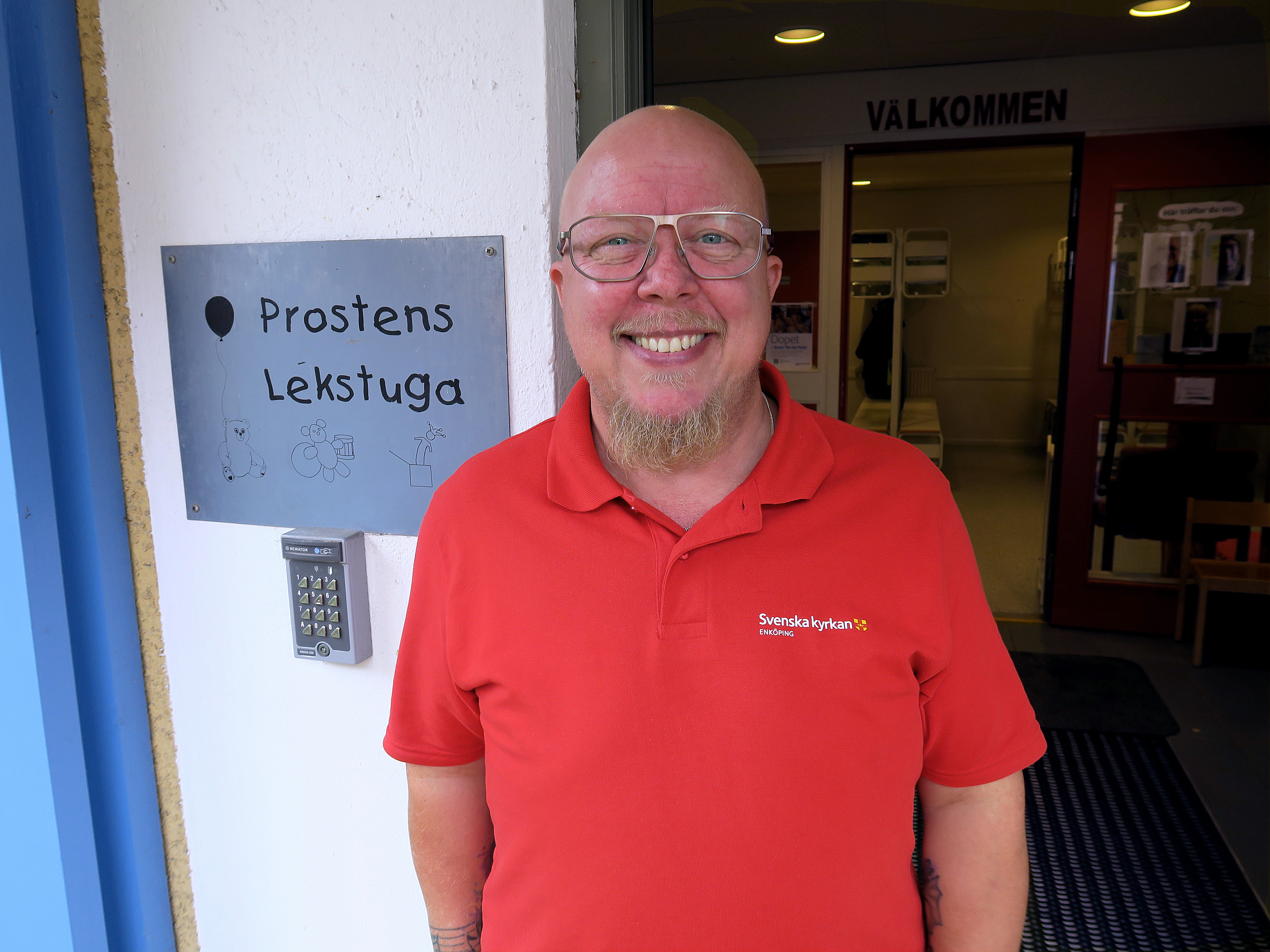 En man med glasögon och röd tröja ler stort mot kameran. På väggen bredvid honom sitter en skylt med texten "Prostens lekstuga".
