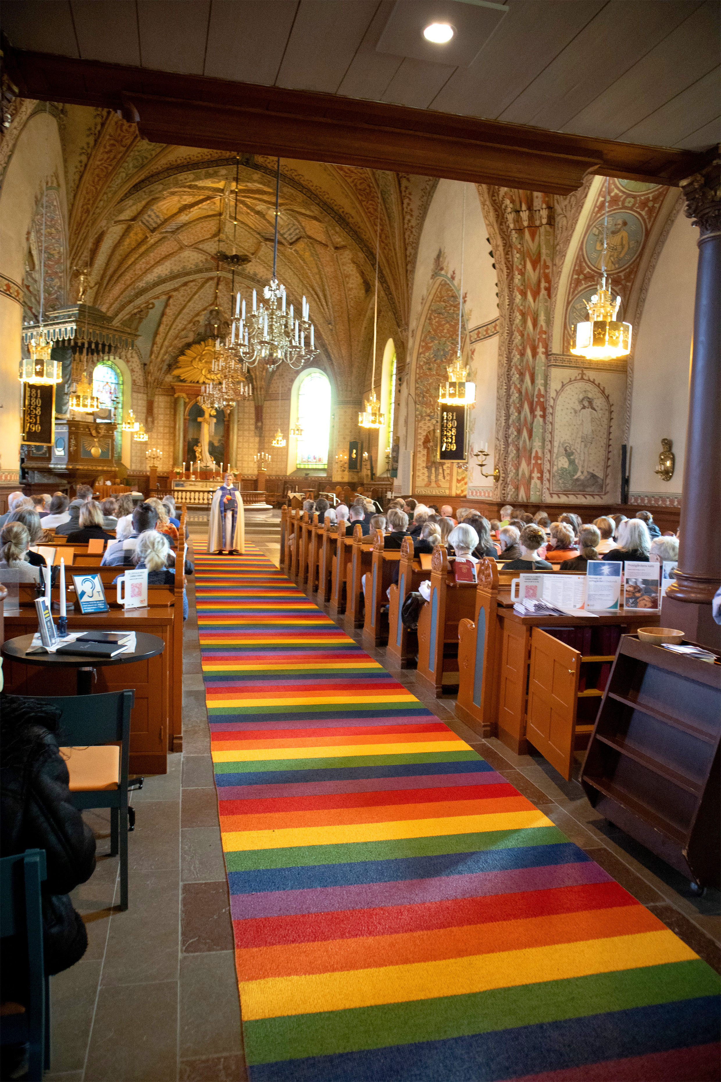 En tvärrandig matta i regnbågens färger ligger i mittgången av en kyrka. I bänkarna sitter folk och i fonden står en präst vänd mot församlingen.