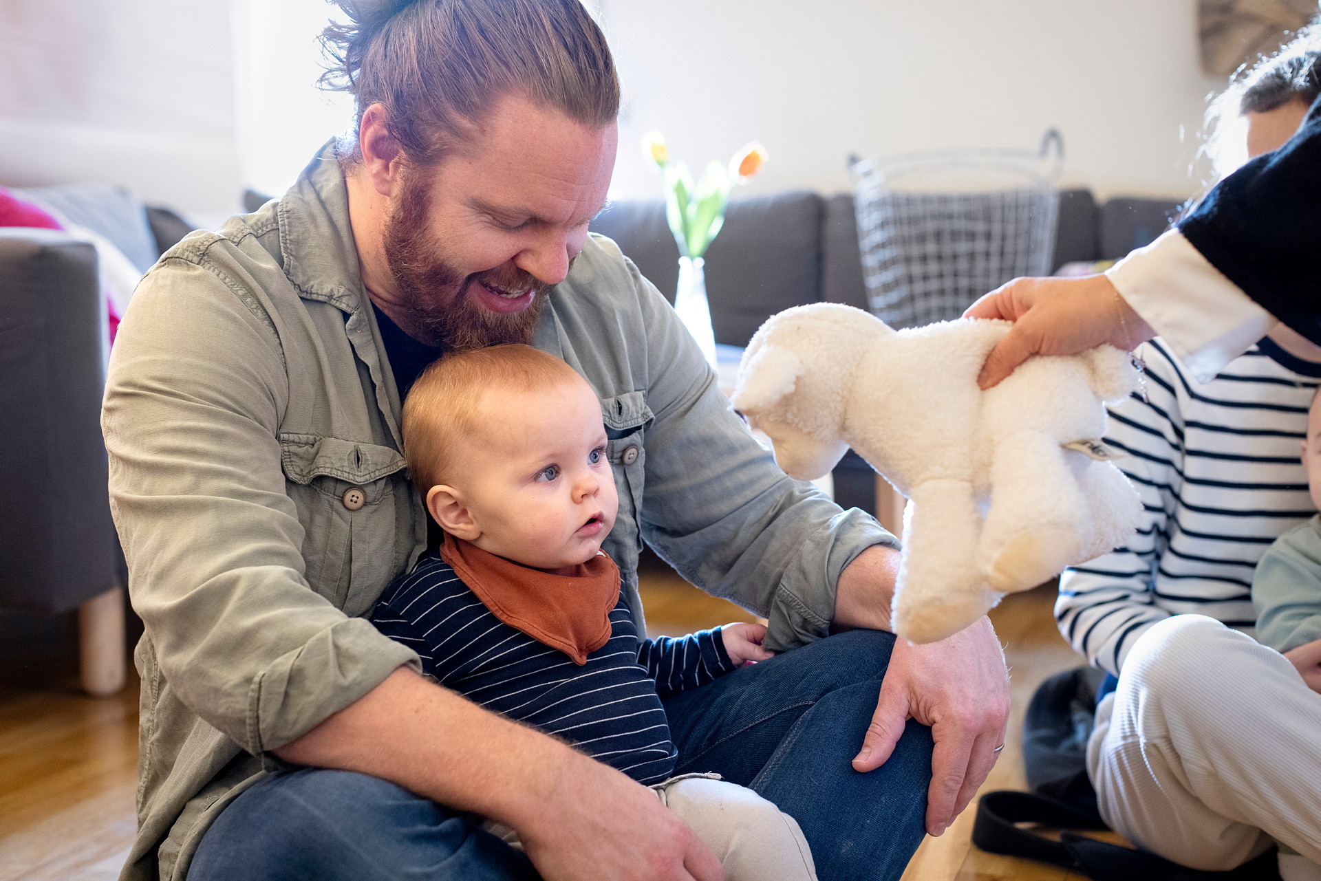 En man i skägg sitter på golvet i skräddarställning med ett litet barn i knät. En person sträcker fram ett gosedjur till barnet.