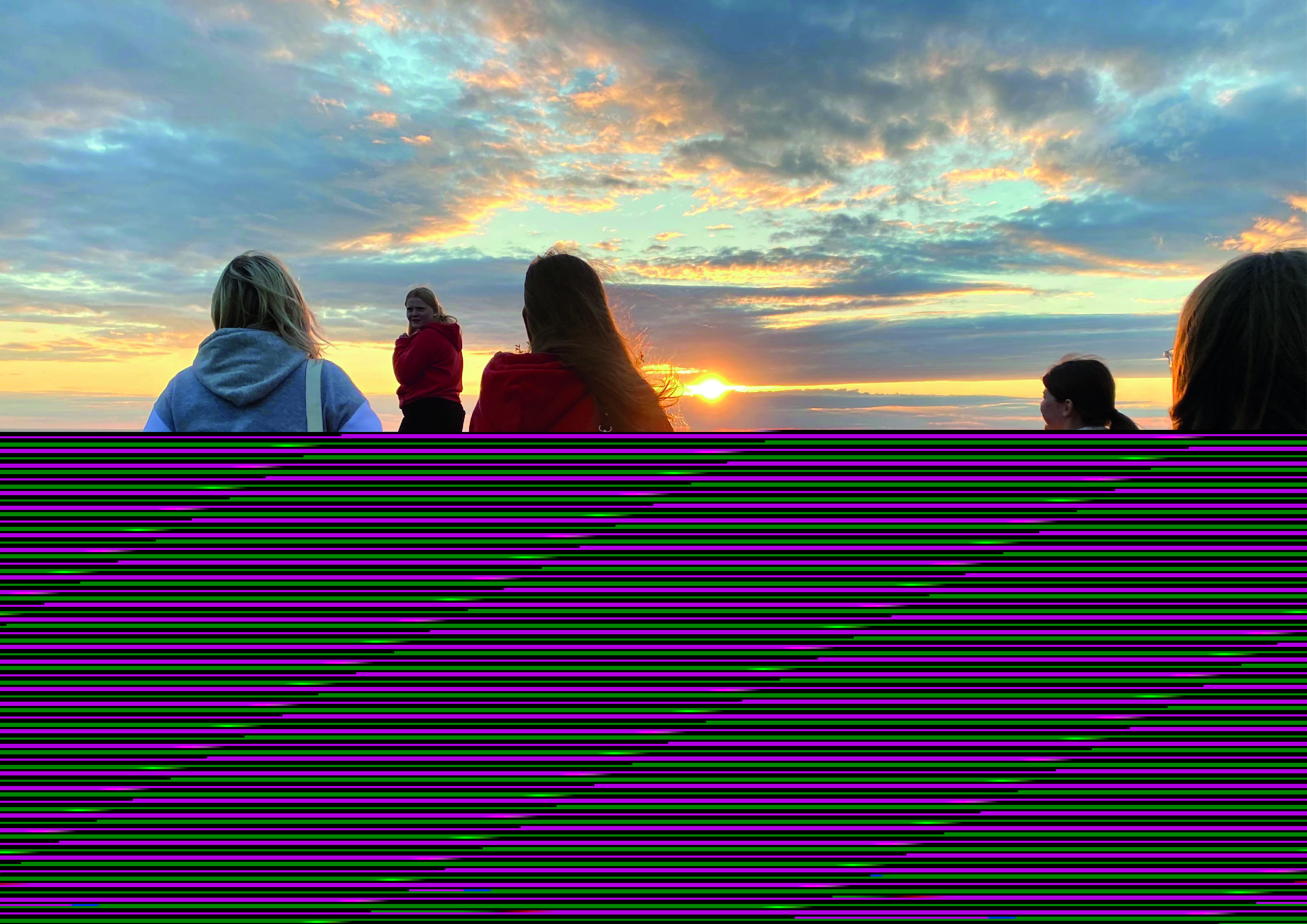 Ungdomar vid en klintkant, blickar ut över solnedgången. Det blåser i deras hår. 