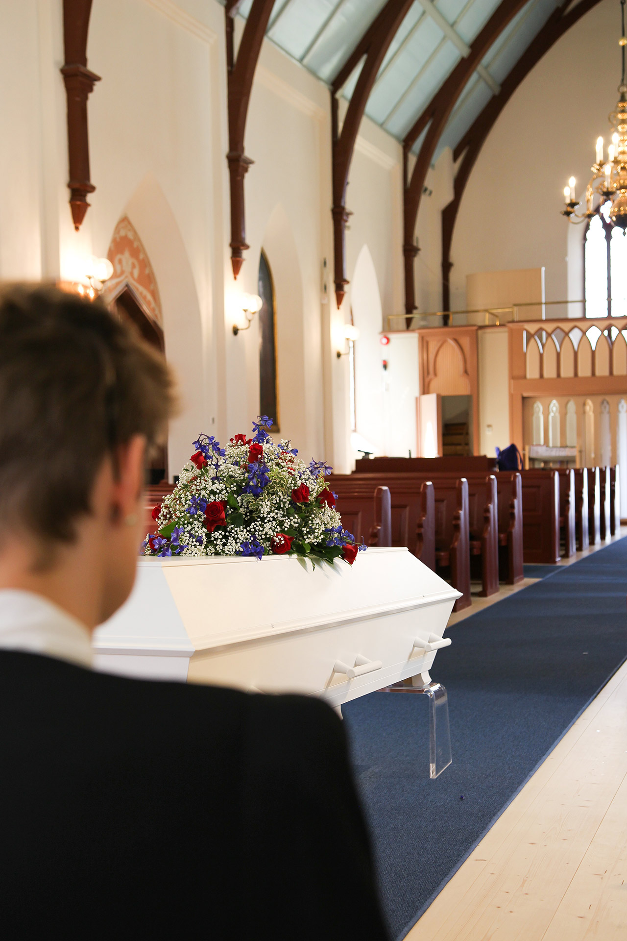 En vit kista med ett stort blomsterarrangemang står i kyrkan. En person i förgrunden kollar på kistan.