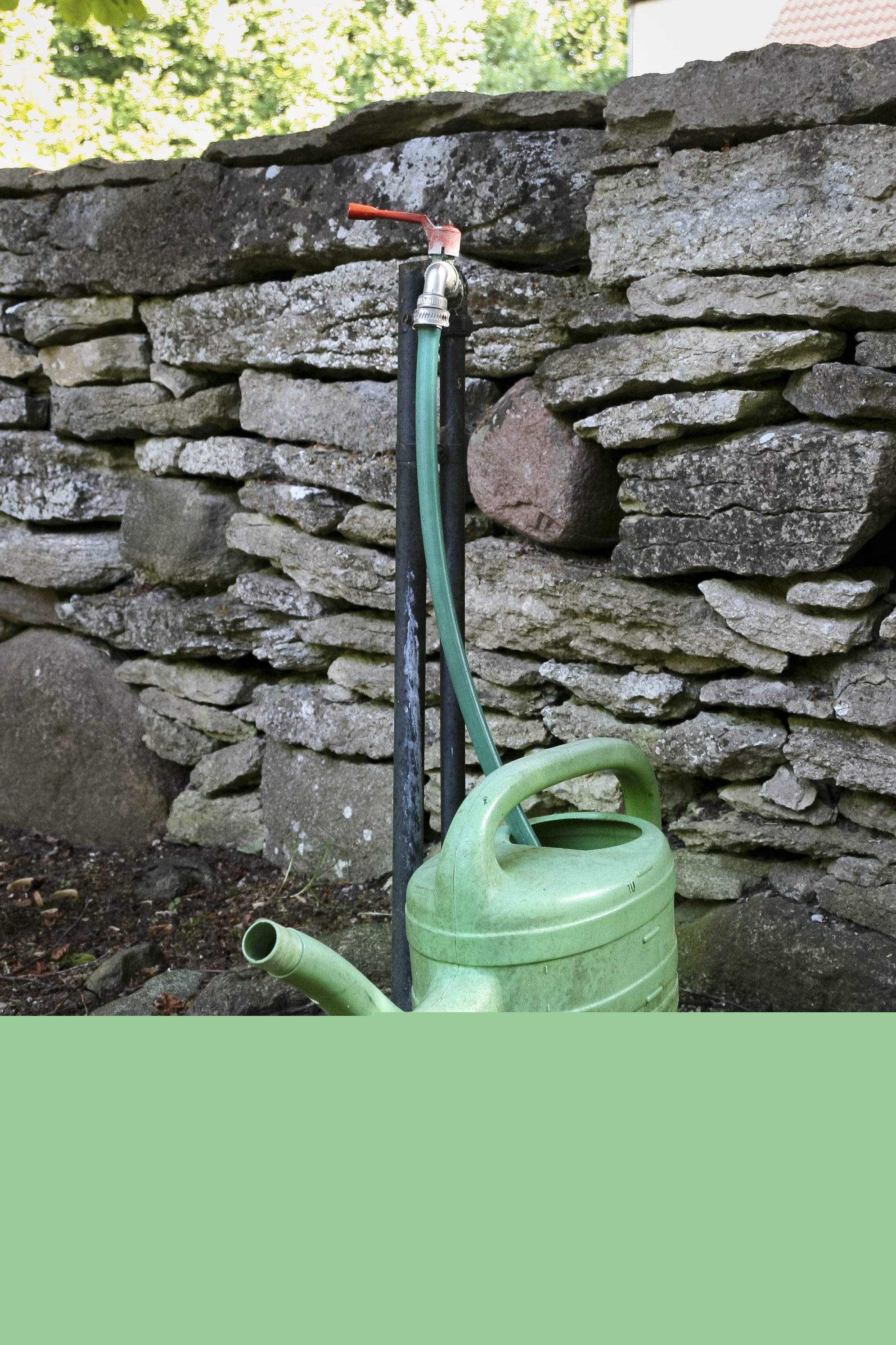 En grön vattenkanna och en vattenslang vid en stenmur.