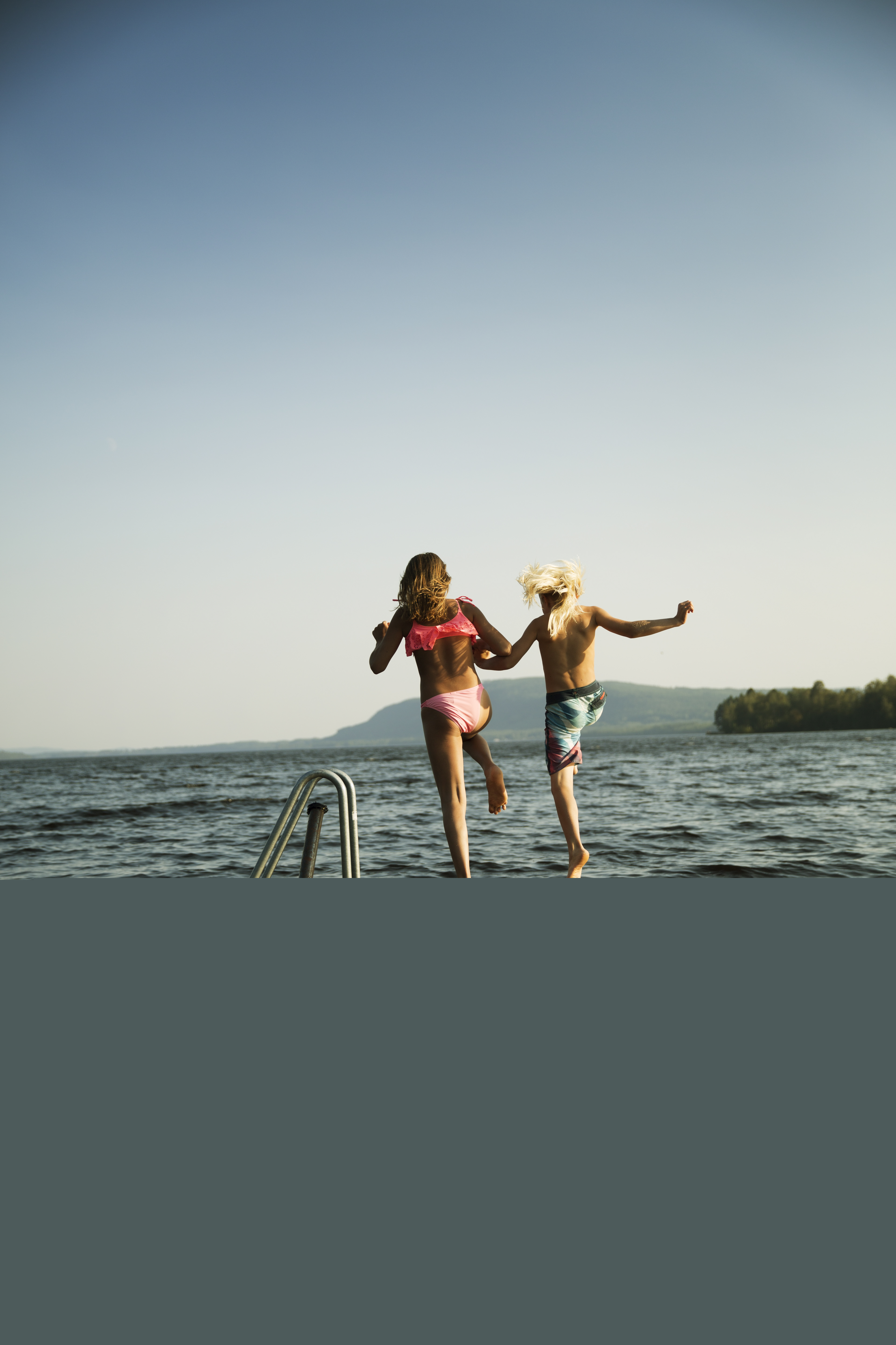 Två barn håller hand och hoppar från bryggan i vattnet.
