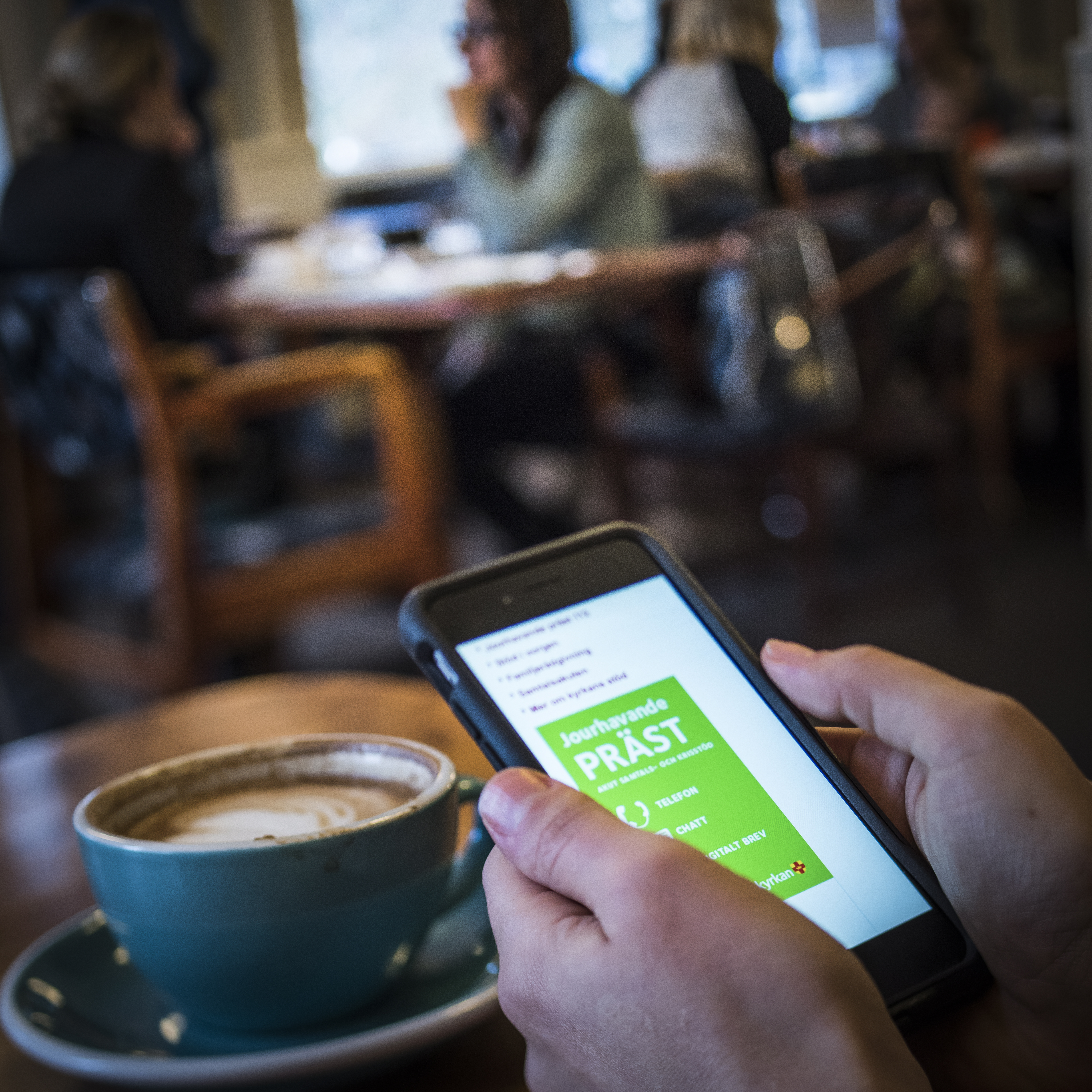 En person som sitter på ett fik har Jourhavande präst uppe på sin mobiltelefon. En kopp kaffe står på bordet.