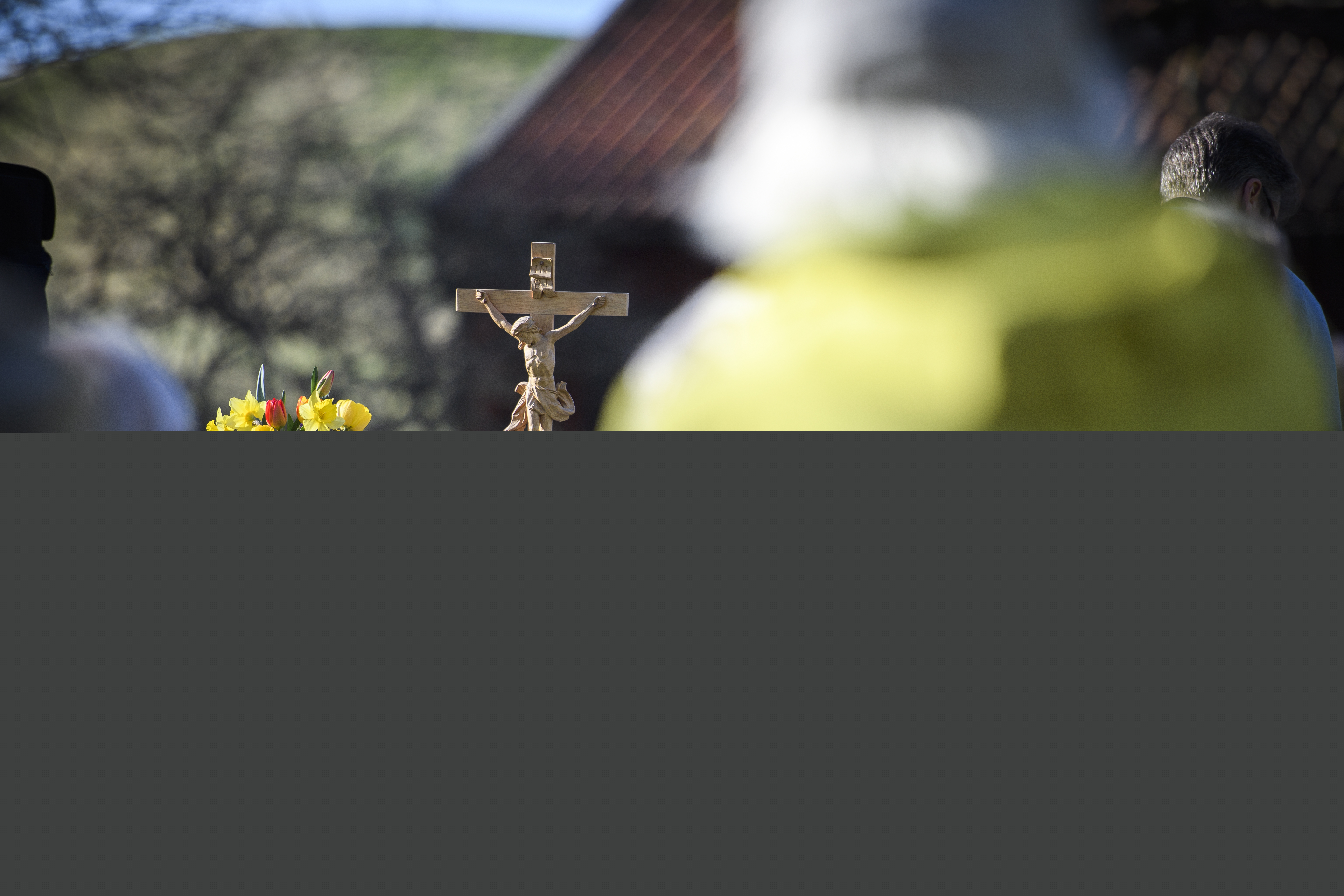 Ett krucifix i trä och en vas med påskblommor står på ett altare utomhus. Suddiga personer i förgrunden.