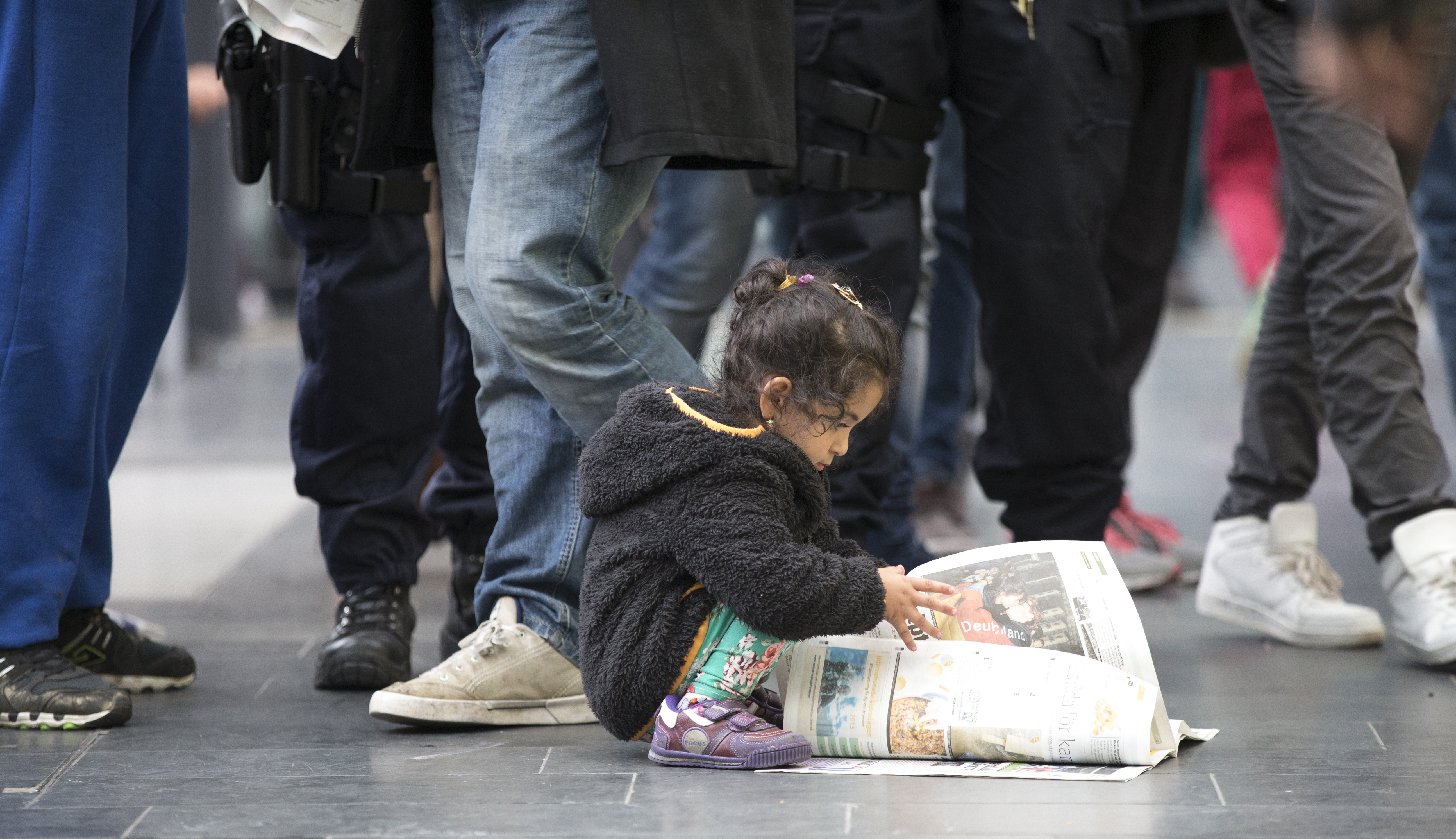 En liten flicka sitter på huk i en folkmassa och bläddrar i en tidning.